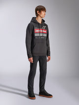 Alpinestars Harken Hoodie, Black pullover fleece hoodie, featuring a large chest graphic with red Alpinestars wordmark and star logo, kangaroo pocket, worn by a model with black pants and sneakers, casual motorsport streetwear