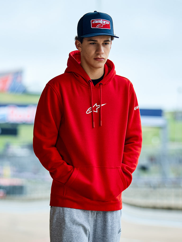 Alpinestars Frontal Hoodie, czerwona polarowa bluza z kapturem wkładana przez głowę, z białym logo Astars na piersi, kieszenią kangurką i kapturem ze sznurkiem, noszona przez mężczyznę w niebieskiej czapce, codzienna odzież streetwearowa w stylu motorsport