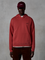 Alpinestars Essentials Ageless Hoodie, casualowa bluza streetwear, kolor ceglasty, małe czarne logo gwiazdy na piersi, kieszeń typu kangurka, wykończenie stone-wash, model w bordowej czapce i brązowych spodniach sztruksowych