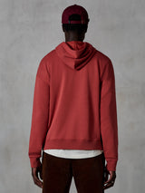 Alpinestars Essentials Ageless Hoodie, casualowa bluza z kapturem, kolor ceglasty, widok od tyłu na modela ubranego w odzież lifestylową, prosty design z prążkowanym dołem i mankietami, stylizacja z brązowymi spodniami sztruksowymi i bordową czapką