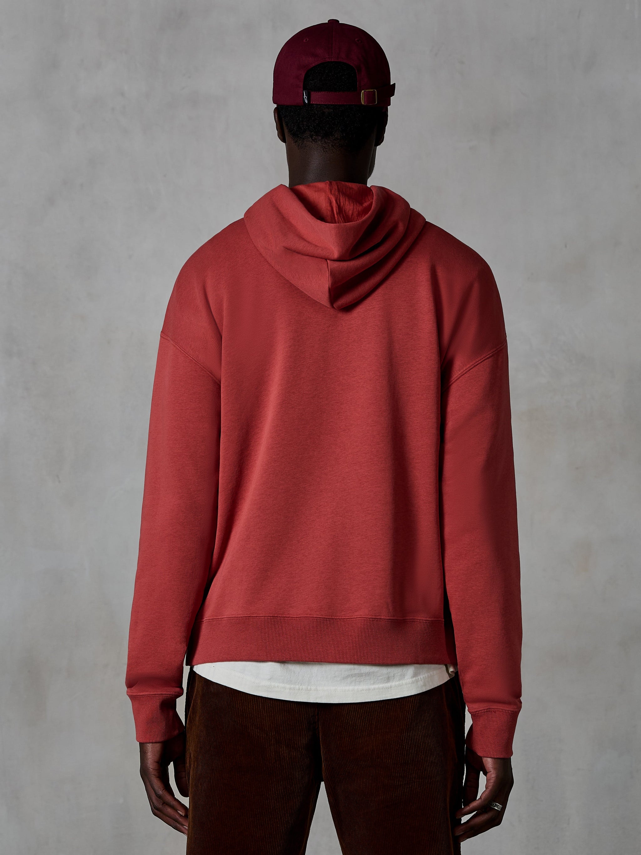 Alpinestars Essentials Ageless Hoodie, casualowa bluza z kapturem, kolor ceglasty, widok od tyłu na modela ubranego w odzież lifestylową, prosty design z prążkowanym dołem i mankietami, stylizacja z brązowymi spodniami sztruksowymi i bordową czapką