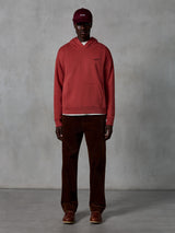 Alpinestars Essentials Ageless Hoodie, sportowa bluza z kapturem w kolorze ceglastym z wykończeniem stone wash, z małym czarnym logo Alpinestars na piersi i przednią kieszenią typu kangurka, noszona przez modela w bordowej czapce i brązowych sztruksowych spodniach, czysty streetwearowy styl.