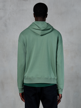 Alpinestars Essentials Ageless Hoodie, codzienna odzież sportowa, kolor zielony (Cactus), widok z tyłu na modelu, wykończenie typu stone wash, zestawiona z czarnymi spodniami, minimalistyczny lifestylowy design