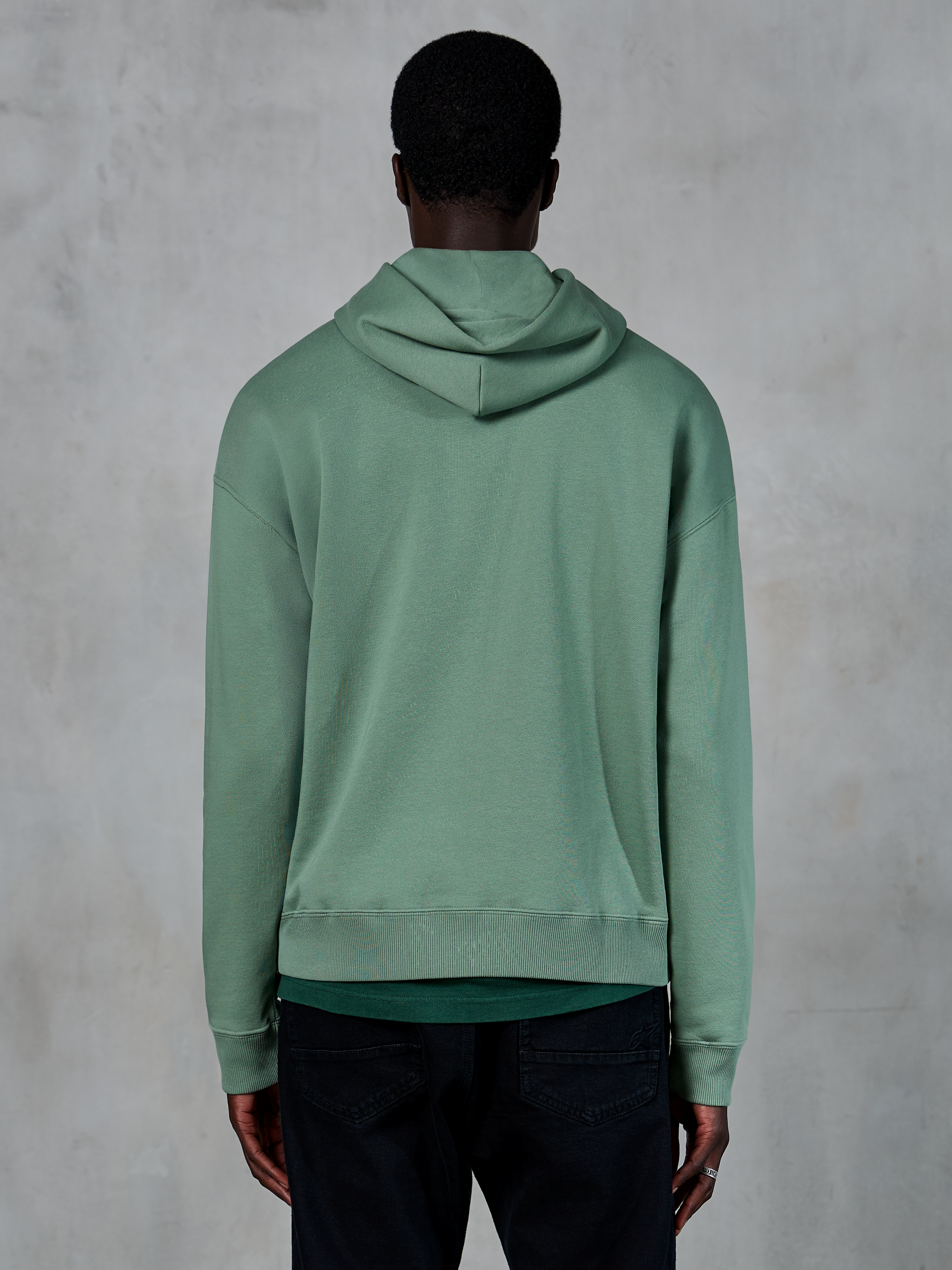 Alpinestars Essentials Ageless Hoodie, codzienna odzież sportowa, kolor zielony (Cactus), widok z tyłu na modelu, wykończenie typu stone wash, zestawiona z czarnymi spodniami, minimalistyczny lifestylowy design