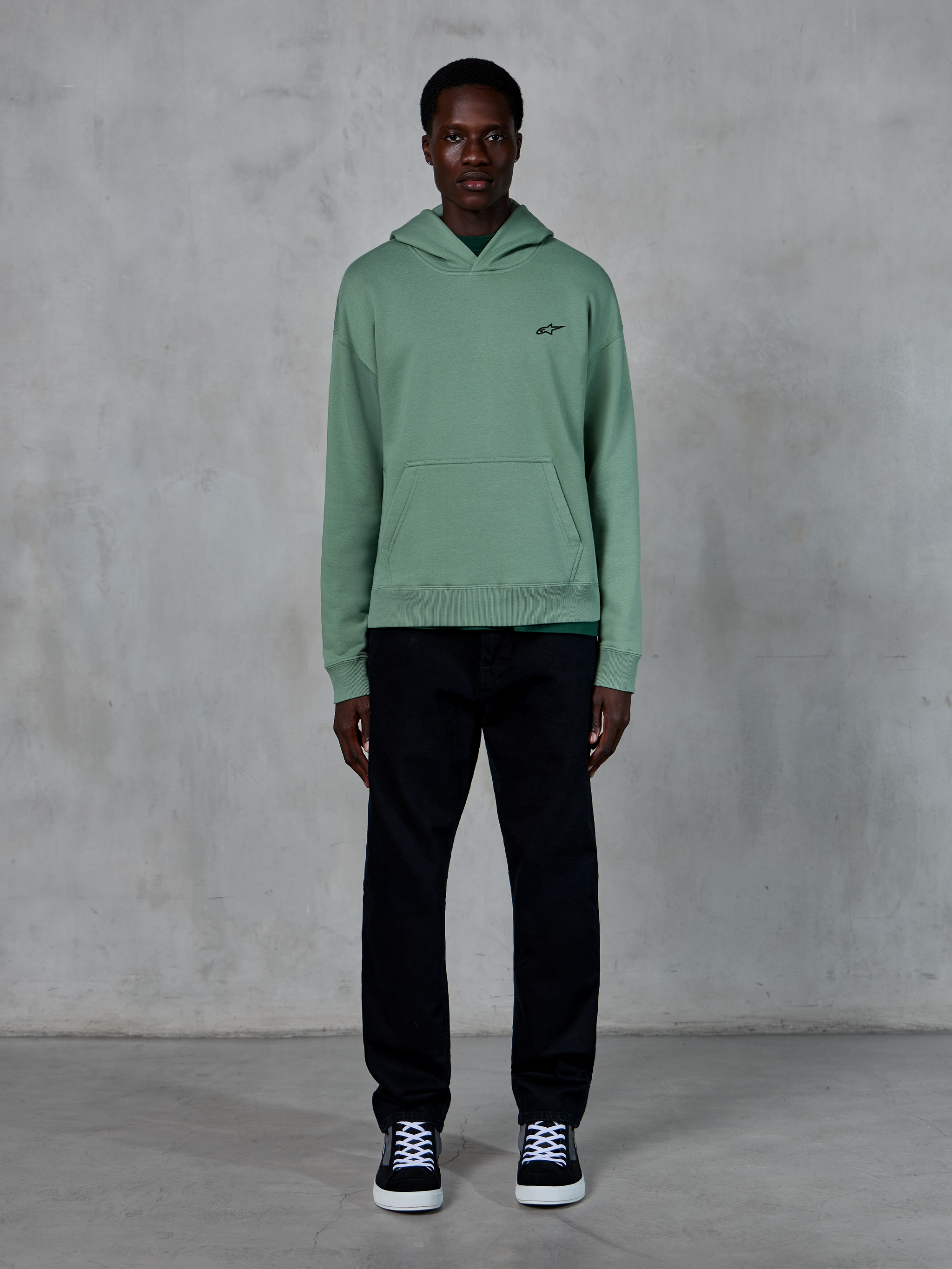 Bluza z kapturem Alpinestars Essentials Ageless, kolor zielony Cactus, małe czarne logo na piersi, kieszeń typu kangurka, casualowa odzież streetwearowa noszona z czarnymi spodniami i czarno-białymi sneakersami na tle teksturowanej ściany