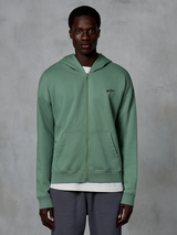 Alpinestars Essentials Ageless Zip Hoodie, casualowa bluza streetwearowa na zamek, kolor zielony Cactus, z subtelnym haftowanym logo na piersi, dzielonymi kieszeniami typu kangurka i ściągaczami, noszona przez modela na teksturowanym szarym tle