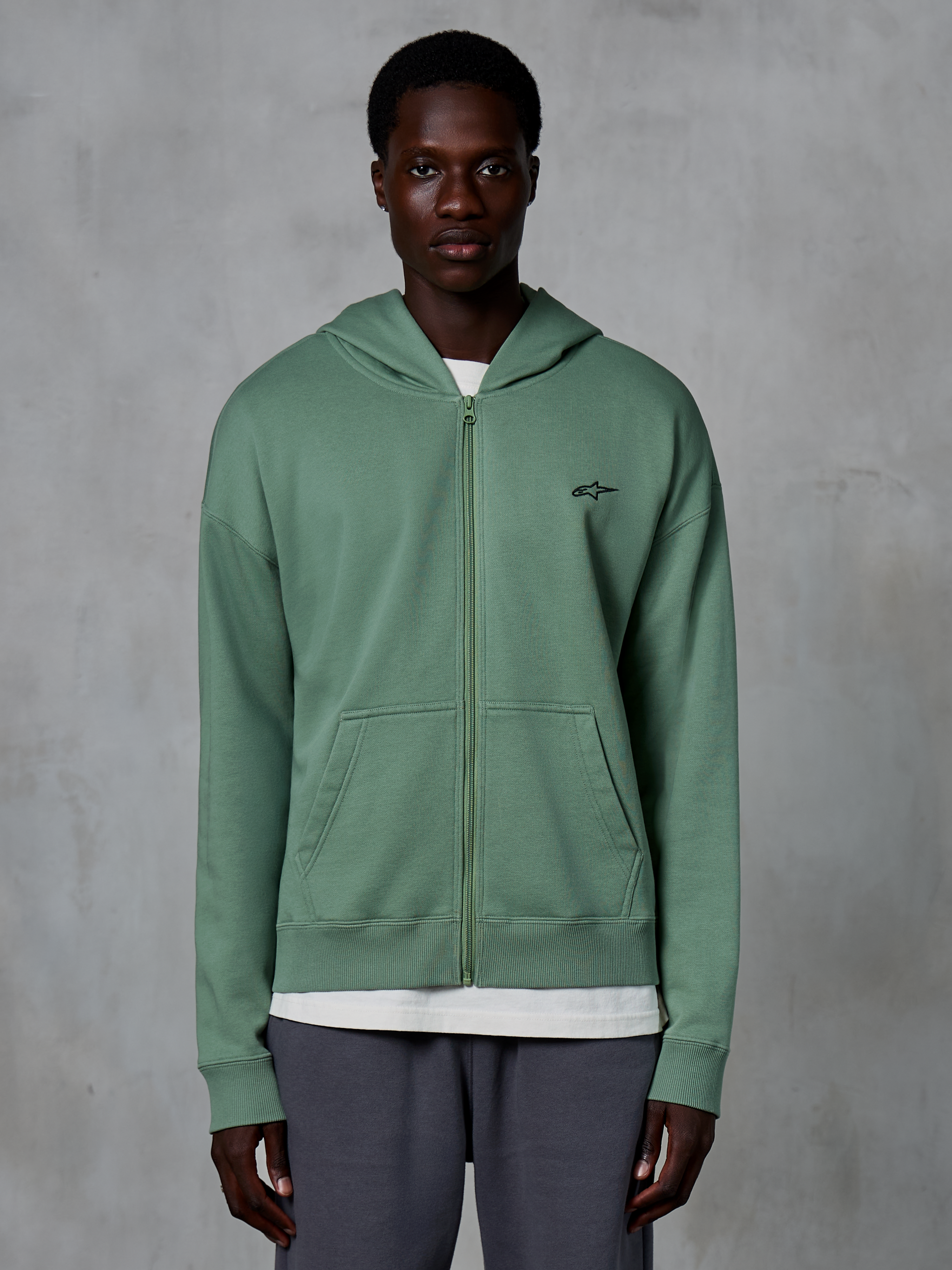 Alpinestars Essentials Ageless Zip Hoodie, casualowa bluza streetwearowa na zamek, kolor zielony Cactus, z subtelnym haftowanym logo na piersi, dzielonymi kieszeniami typu kangurka i ściągaczami, noszona przez modela na teksturowanym szarym tle