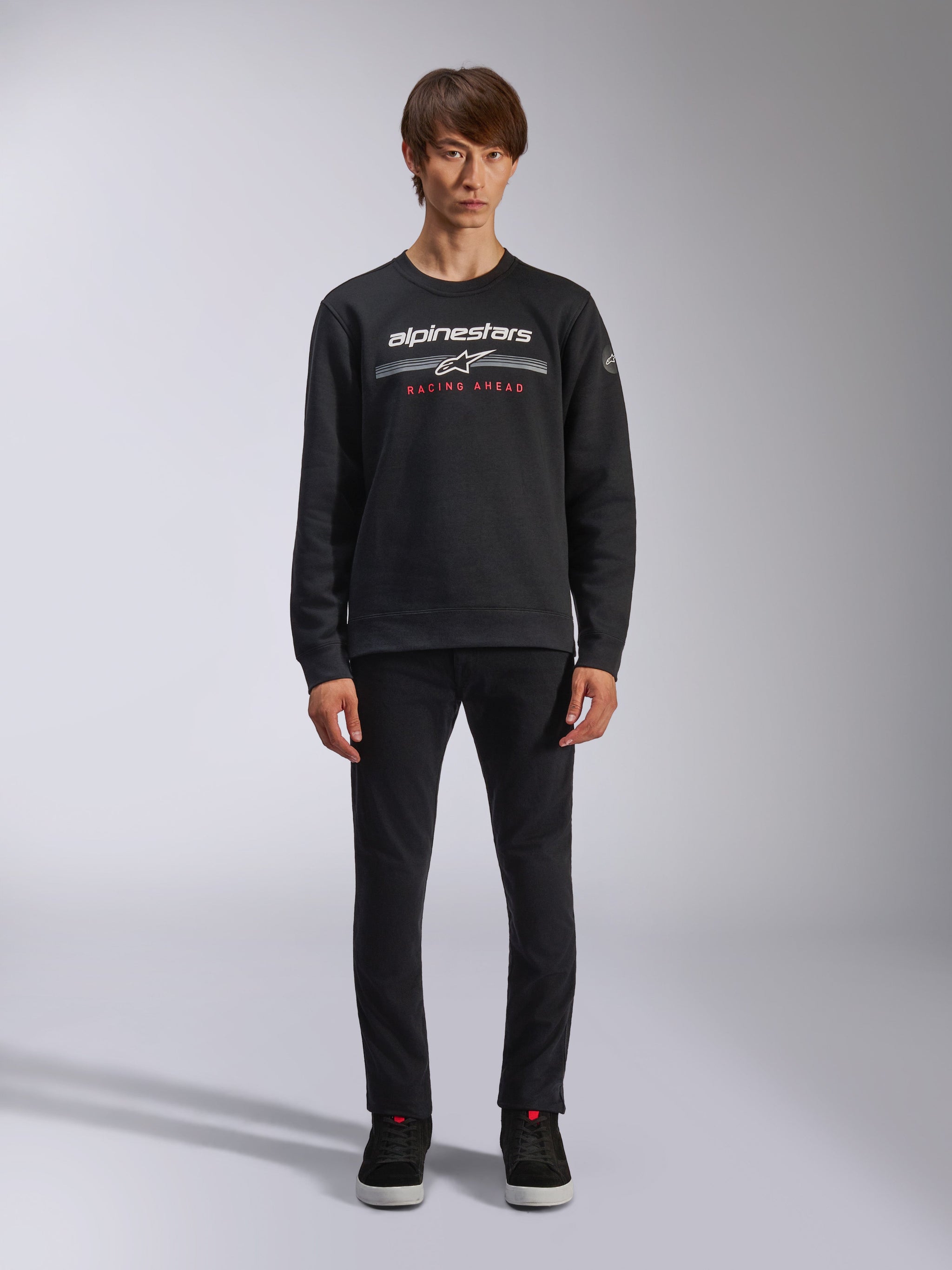 Alpinestars Bettern Crew, czarna bluza typu crew neck, białe logo Alpinestars z szarymi poziomymi pasami i czerwonym napisem „RACING AHEAD”, codzienna odzież motorsportowa w stylu streetwear, prezentowana na modelu z czarnymi spodniami i tenisówkami