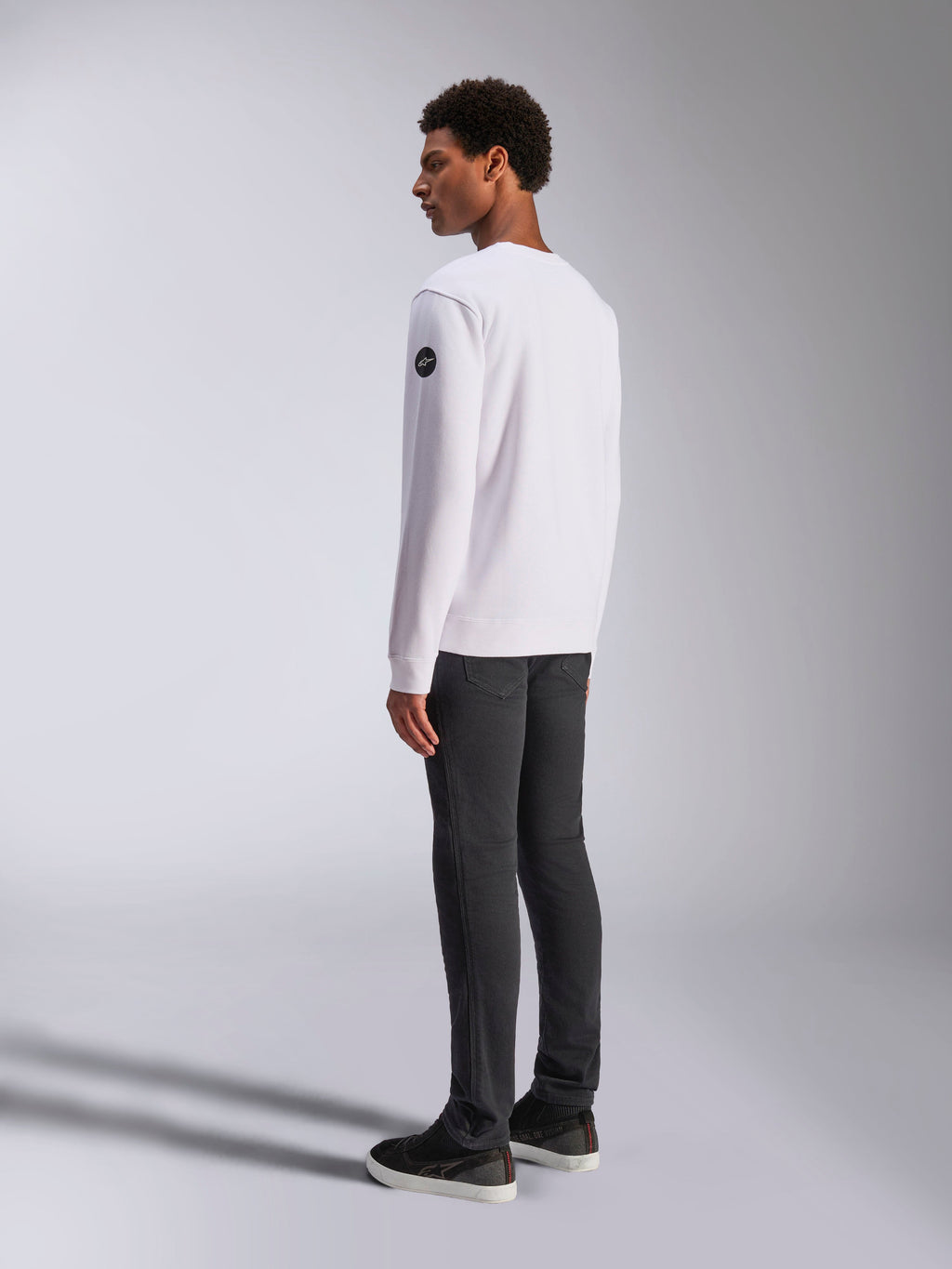 Alpinestars Bettern Crew, biała bluza typu crewneck, widok z tyłu i z boku, czarne okrągłe logo na rękawie, zestawiona z ciemnoszarymi spodniami i czarnymi sneakersami, casualowy motorsportowy styl streetwear