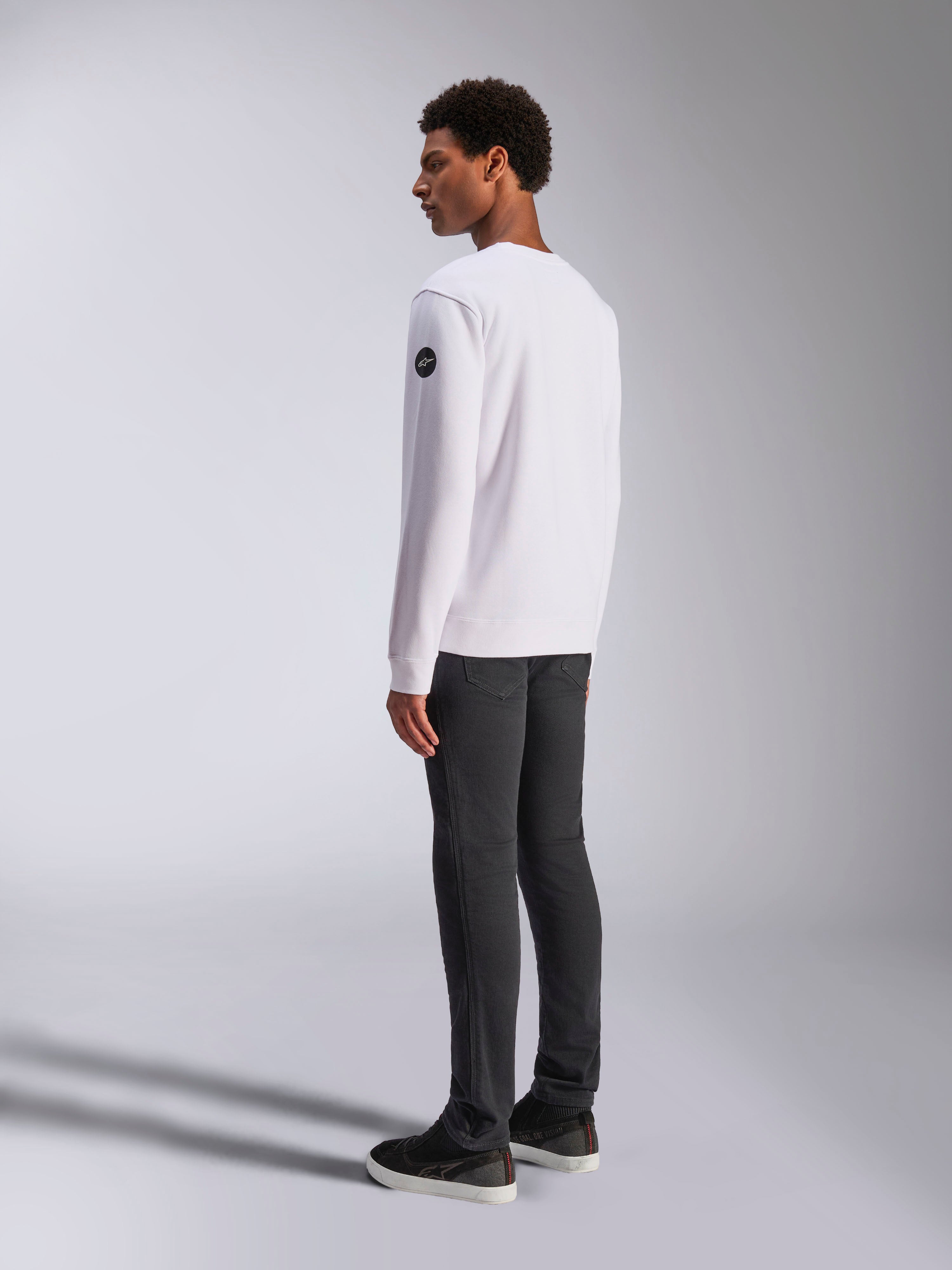Alpinestars Bettern Crew, biała bluza typu crewneck, widok z tyłu i z boku, czarne okrągłe logo na rękawie, zestawiona z ciemnoszarymi spodniami i czarnymi sneakersami, casualowy motorsportowy styl streetwear