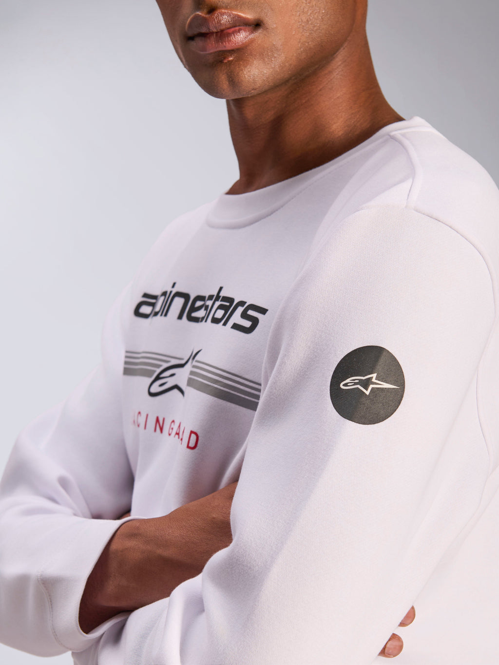 Alpinestars Bettern Crew, biała bluza typu crewneck, codzienna odzież motorsportowa typu streetwear, z grafiką na piersi z logotypem Alpinestars i szarymi paskami, czarna okrągła naszywka z logo gwiazdy na rękawie, polarowa konstrukcja typu pullover