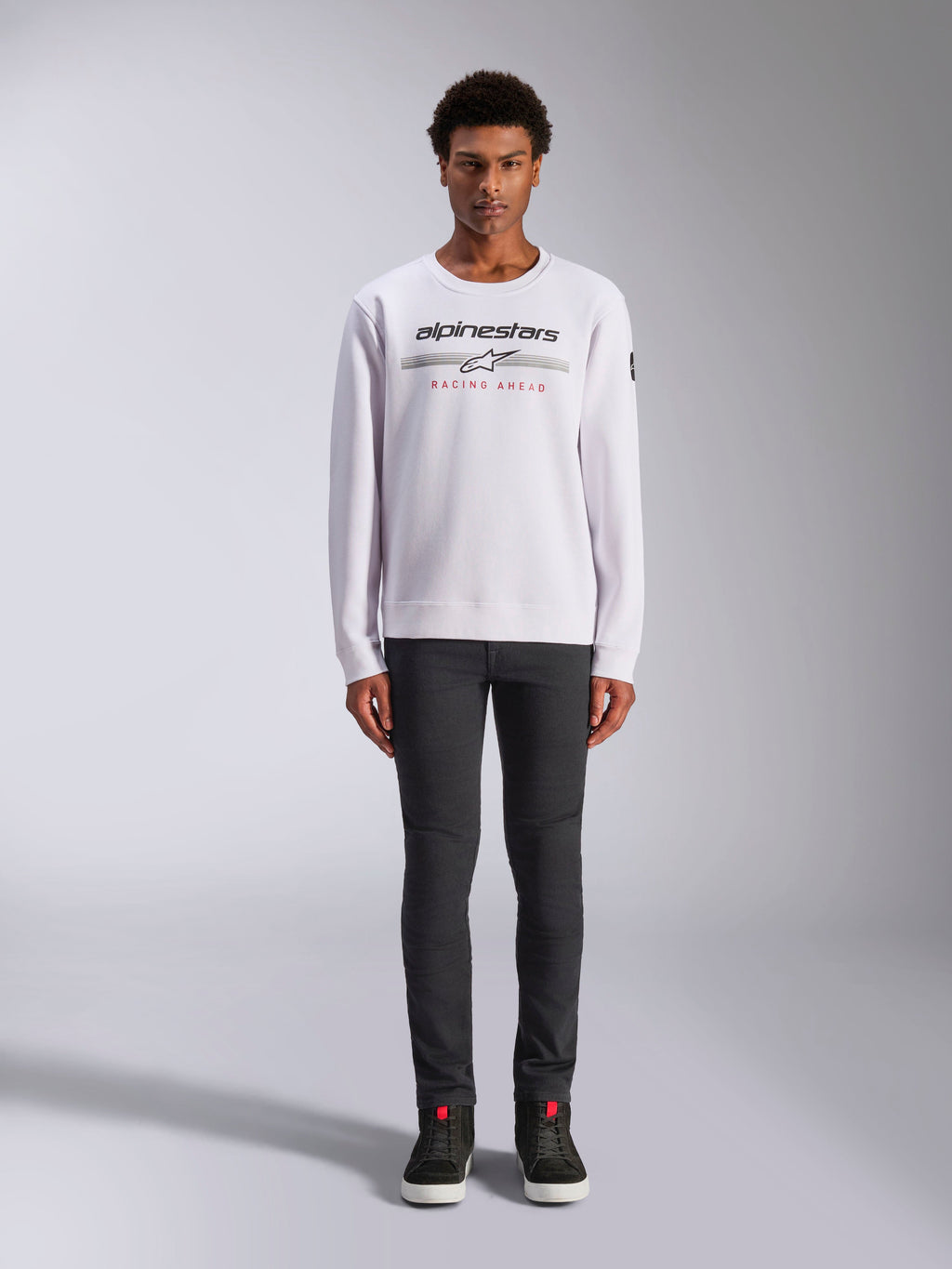 Alpinestars Bettern Crew, biała bluza typu crew neck, model w ciemnoszarych spodniach i czarnych wysokich sneakersach, logo Alpinestars z czerwonym napisem „Racing Ahead”, codzienna odzież uliczna inspirowana sportami motorowymi