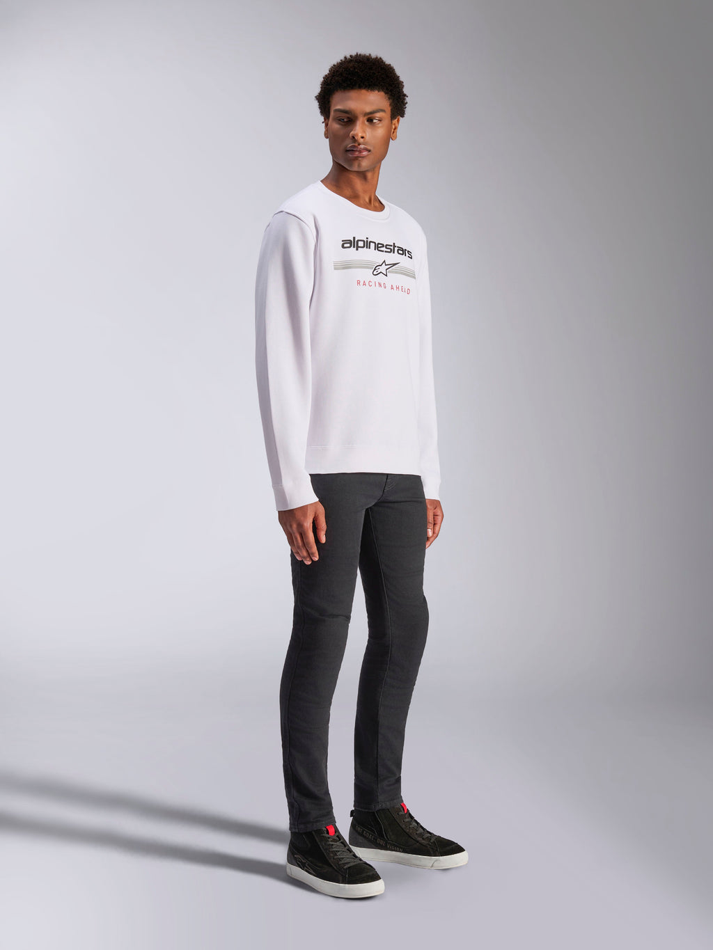 Alpinestars Bettern Crew, biała bluza typu crewneck z czarnym logo Alpinestars i czerwonym napisem „Racing Ahead”, casualowa odzież inspirowana motorsportem, prezentowana przez modela w ciemnych spodniach i czarnych sneakersach