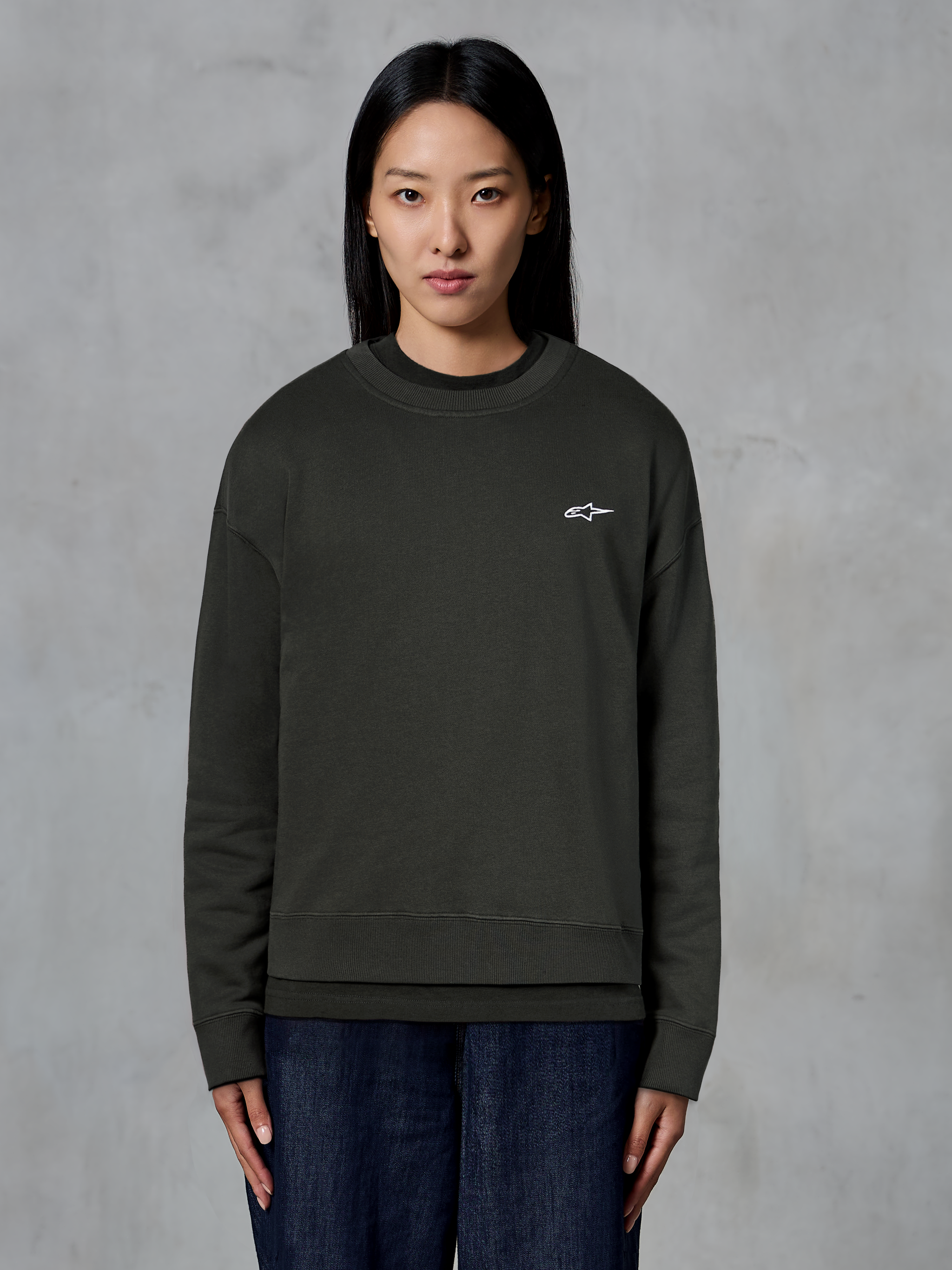 Alpinestars Essentials Ageless Crew, wysokiej jakości bluza z okrągłym dekoltem, kolor Off Black, małe białe logo Alpinestars na piersi, swobodny styl streetwearowy, modelka w ciemnych jeansach