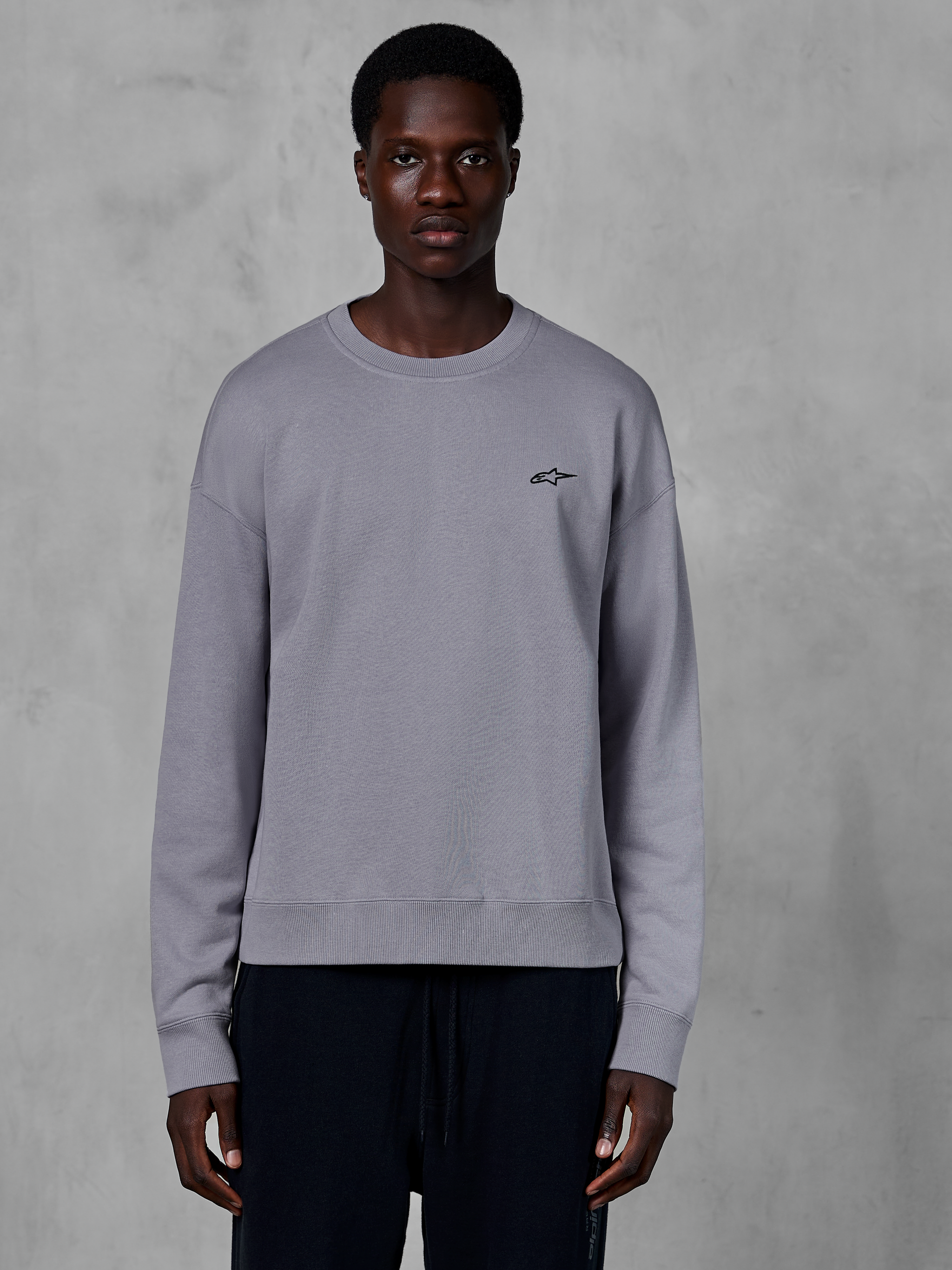 Alpinestars Essentials Ageless Crew, srebrna bluza z okrągłym dekoltem prezentowana na modelu, z małym czarnym logo gwiazdy na piersi, swobodny krój ze ściągaczami przy mankietach i na dole, codzienna odzież uliczna w stylu motorsport