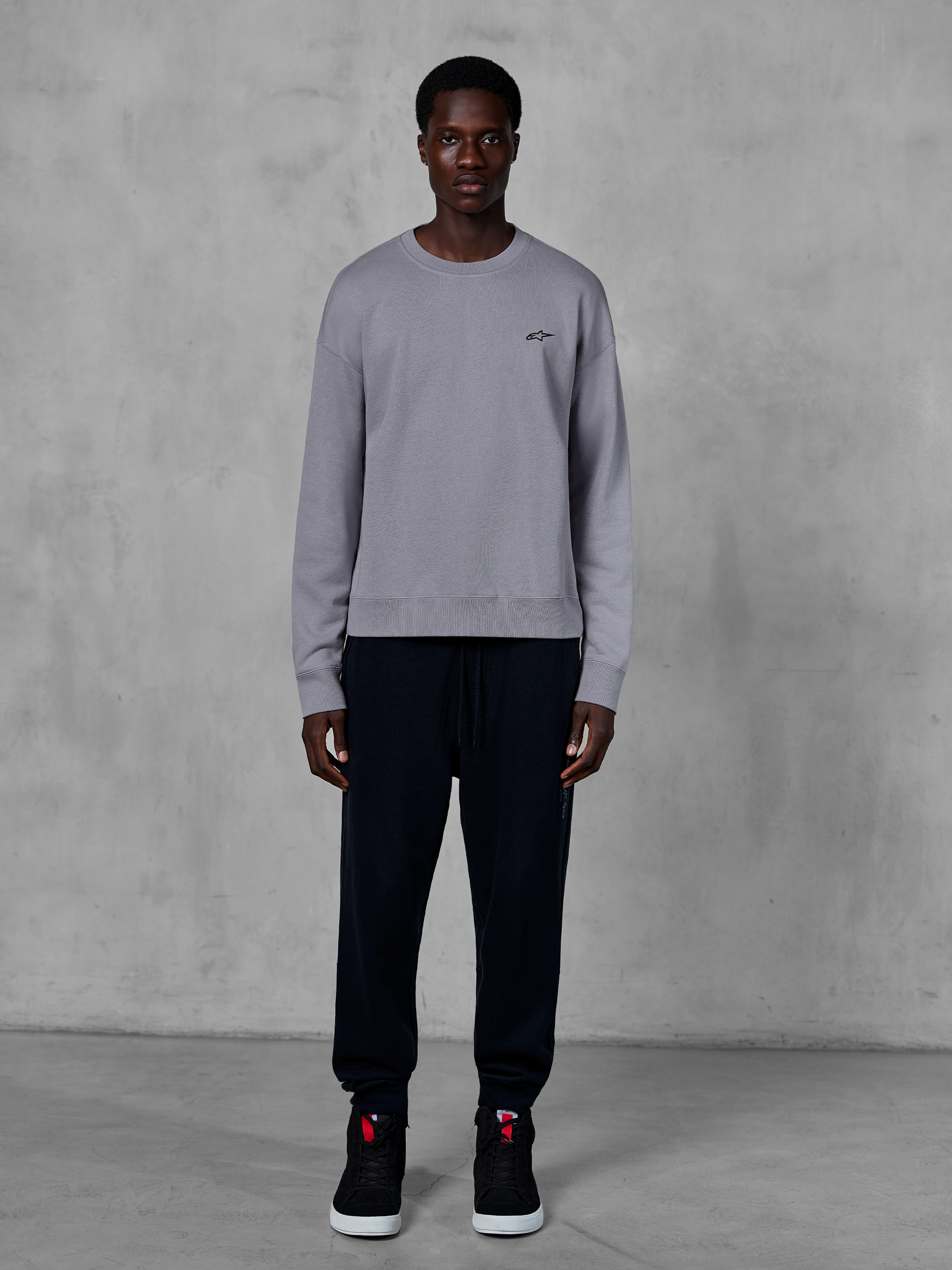 Alpinestars Essentials Ageless Crew, srebrna bluza typu crewneck, prezentowana na modelu z czarnymi joggersami i czarnymi sneakersami, małe czarne logo Alpinestars na piersi, casualowy styl streetwearowy inspirowany motorsportem