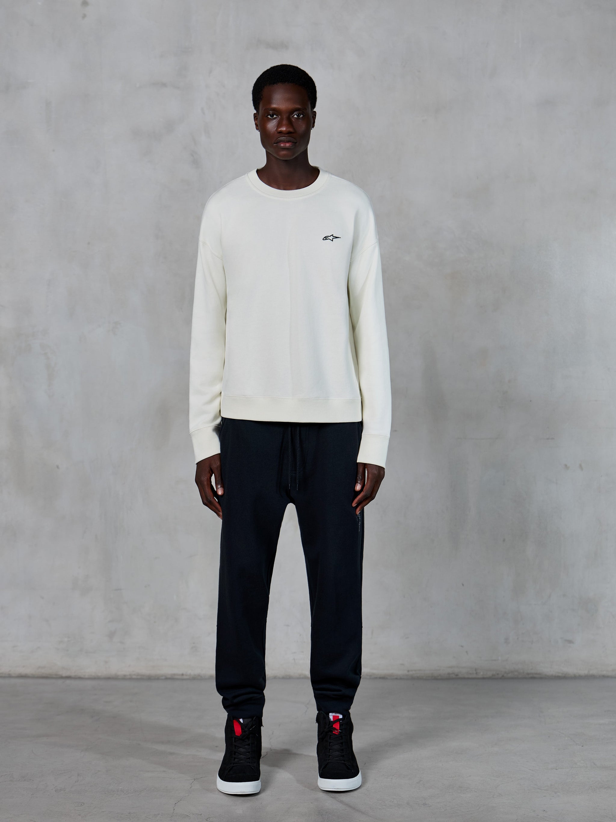 Alpinestars Essentials Ageless Crew, bluza z okrągłym dekoltem, złamana biel z małym czarnym logo gwiazdy, noszona przez modela z czarnymi joggerami, codzienny streetwear inspirowany sportami motorowymi