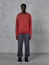 Alpinestars Essentials Ageless Crew, bluza z okrągłym dekoltem w kolorze Brick Red, noszona z szarymi spodniami dresowymi i czarnymi sneakersami, posiada małe czarne logo na piersi, codzienna odzież uliczna inspirowana sportami motorowymi