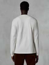 Alpinestars Essentials Ageless LS Tee, koszulka z długim rękawem, złamana biel, widok od tyłu osoby mającej na sobie koszulkę i brązowe sztruksowe spodnie, luźny krój, teksturowana bawełna organiczna, minimalistyczny codzienny styl streetwear