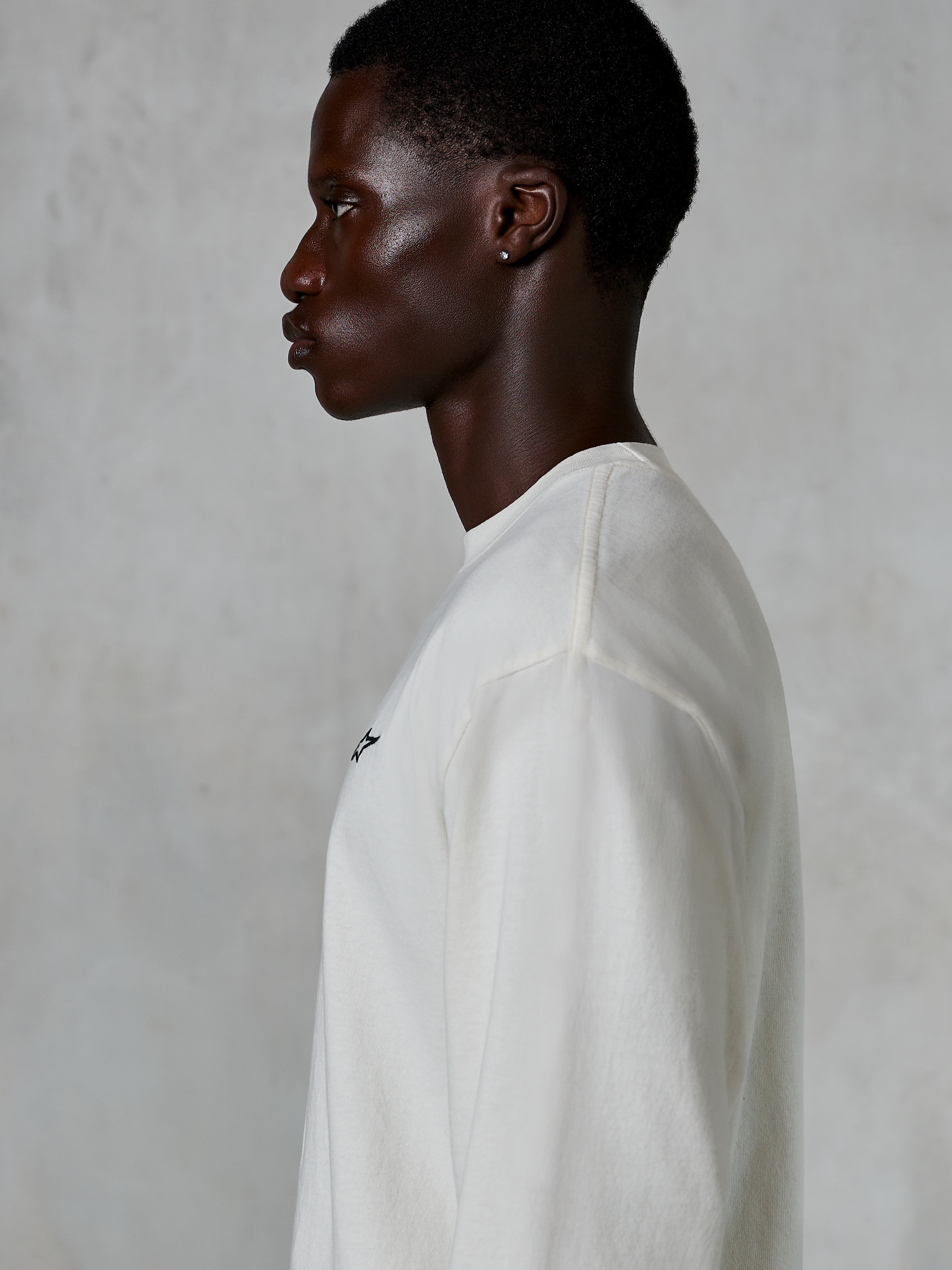 Alpinestars Essentials Ageless LS Tee, koszulka z długim rękawem, kolor Off White, widok z profilu na mężczyznę w koszulce o luźnym kroju, małe czarne logo na piersi, codzienna odzież lifestylowa inspirowana sportami motorowymi