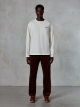 Alpinestars Essentials Ageless LS Tee, koszulka z długim rękawem, kolor off-white, noszona przez modela z brązowymi sztruksowymi spodniami i brązowymi skórzanymi butami, małe czarne logo na piersi, luźny nowoczesny krój, casualowy styl streetwearowy