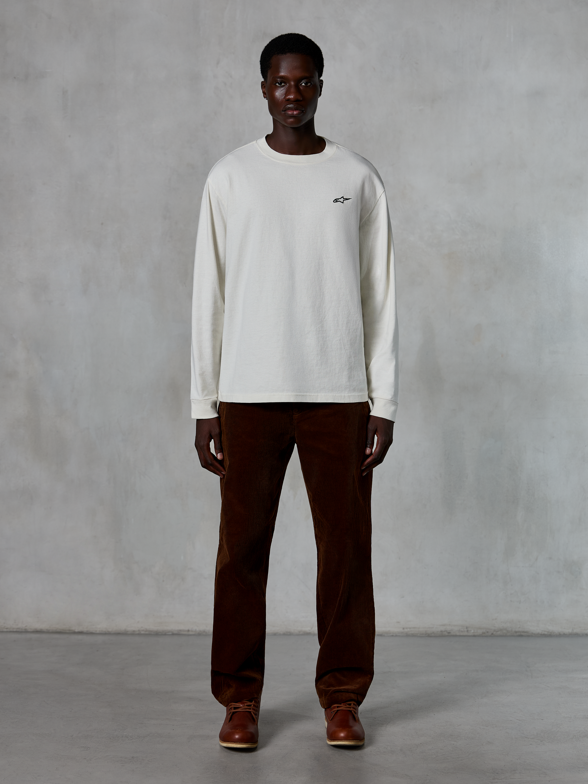 Alpinestars Essentials Ageless LS Tee, koszulka z długim rękawem, kolor off-white, noszona przez modela z brązowymi sztruksowymi spodniami i brązowymi skórzanymi butami, małe czarne logo na piersi, luźny nowoczesny krój, casualowy styl streetwearowy