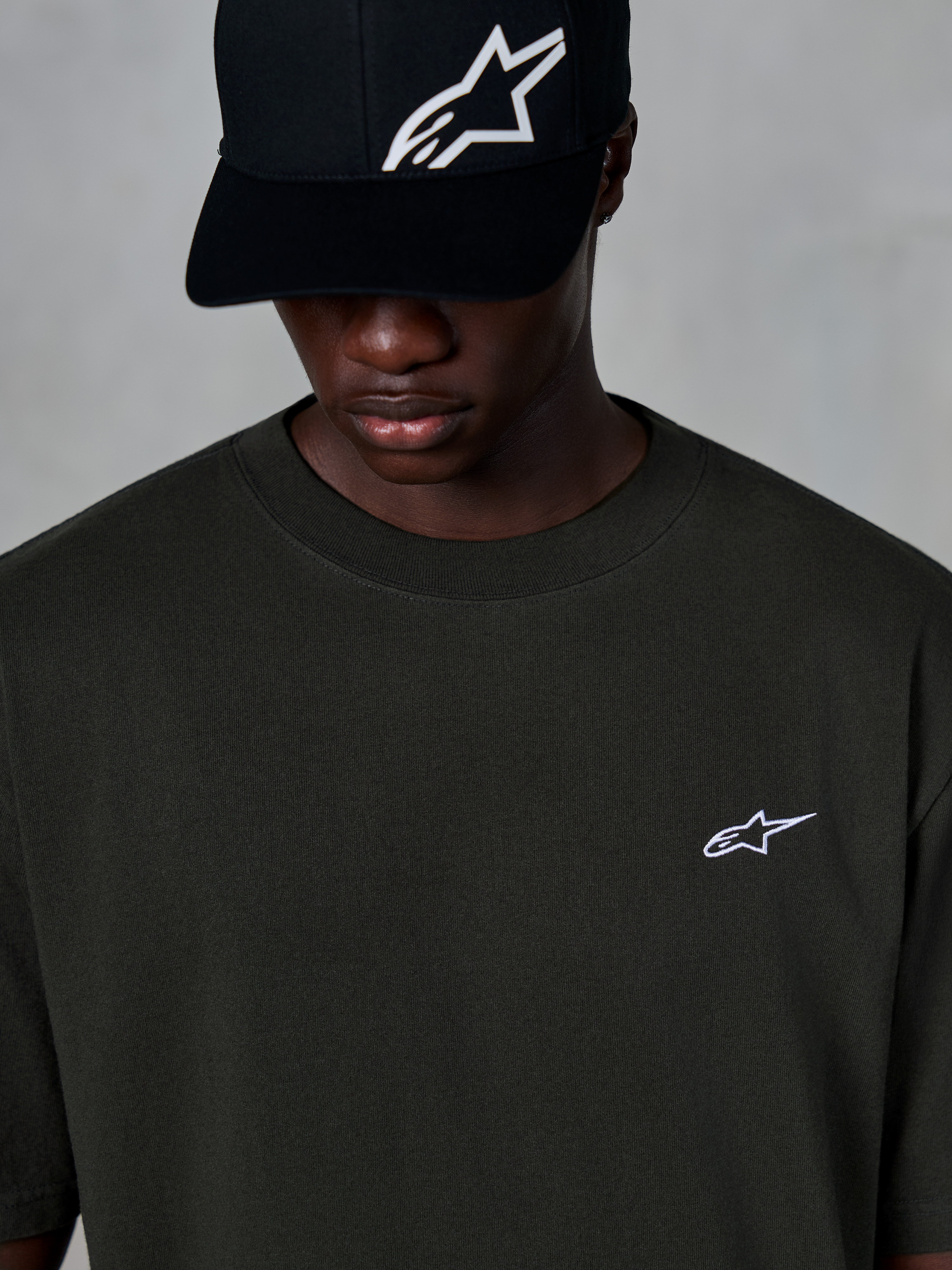 Alpinestars Essentials Ageless SS Tee, koszulka z krótkim rękawem, Off Black, z małym białym logo Alpinestars na piersi, noszona z czarną czapką z daszkiem z logo, casualowy styl streetwearowy inspirowany sportami motorowymi