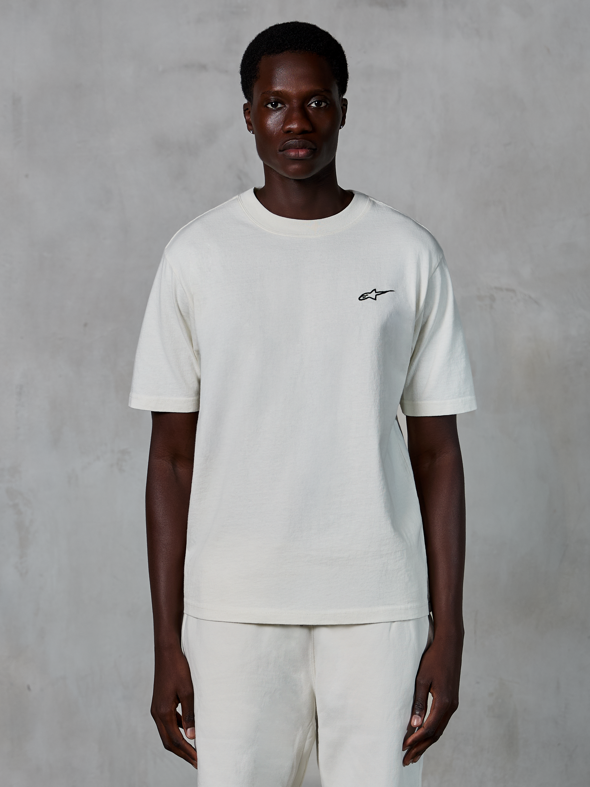 Alpinestars Essentials Ageless SS Tee, koszulka z krótkim rękawem, kolor off-white, prezentowana na modelu, małe czarne logo Alpinestars na piersi, luźny krój, dżersej z bawełny organicznej, casualowy motorsportowy streetwear