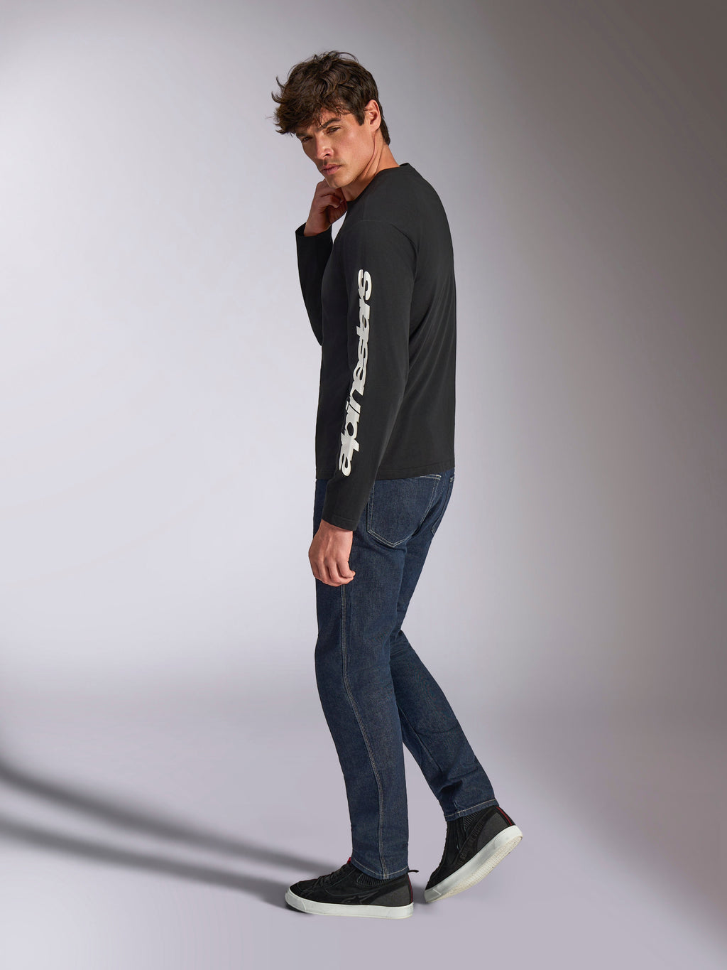 Alpinestars Trackside CSF Long Sleeve Tee, czarna koszulka z długim rękawem z białym pionowym napisem Alpinestars na rękawie, model ubrany w ciemne jeansy i sneakersy, codzienna odzież sportowa inspirowana motorsportem