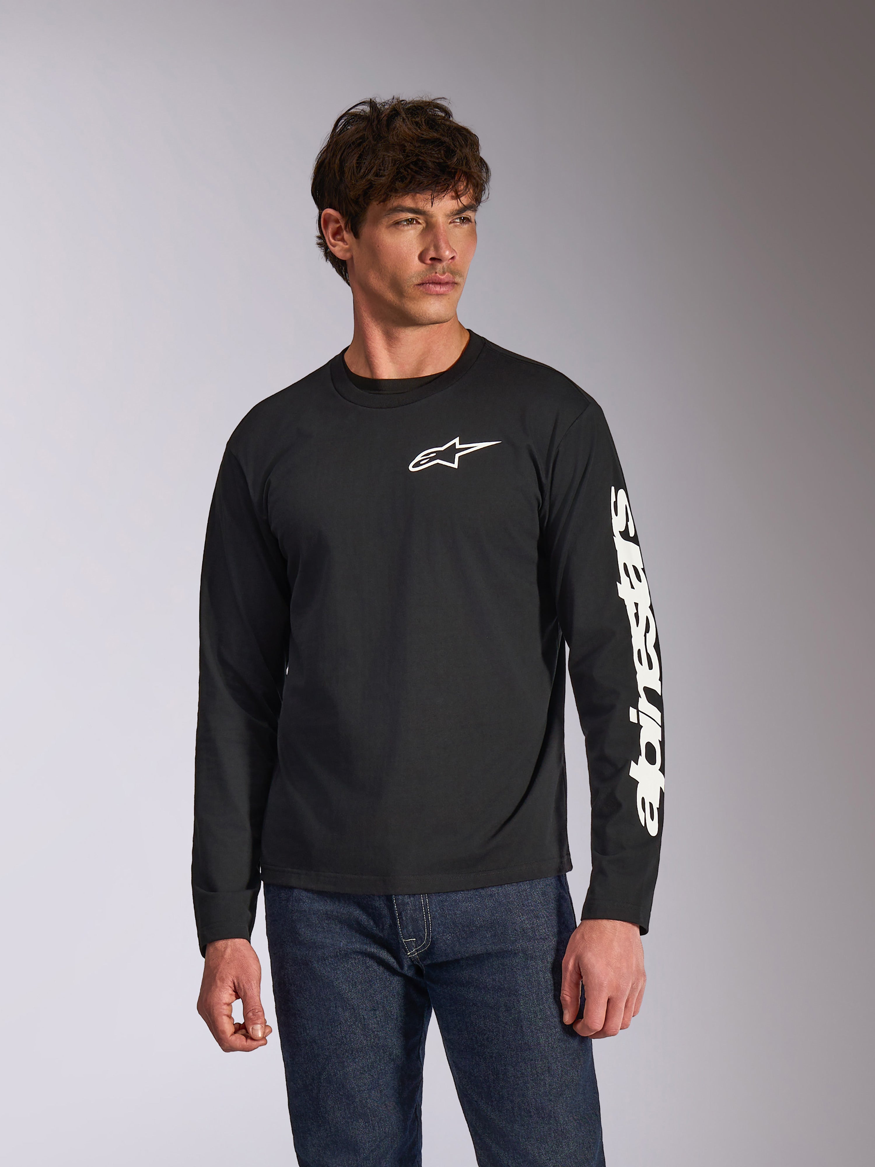 Alpinestars Trackside CSF Long Sleeve Tee, czarna, białe logo Alpinestars na piersi i branding na rękawie, prezentowana przez modela w niebieskich jeansach, codzienna odzież uliczna w stylu motorsport
