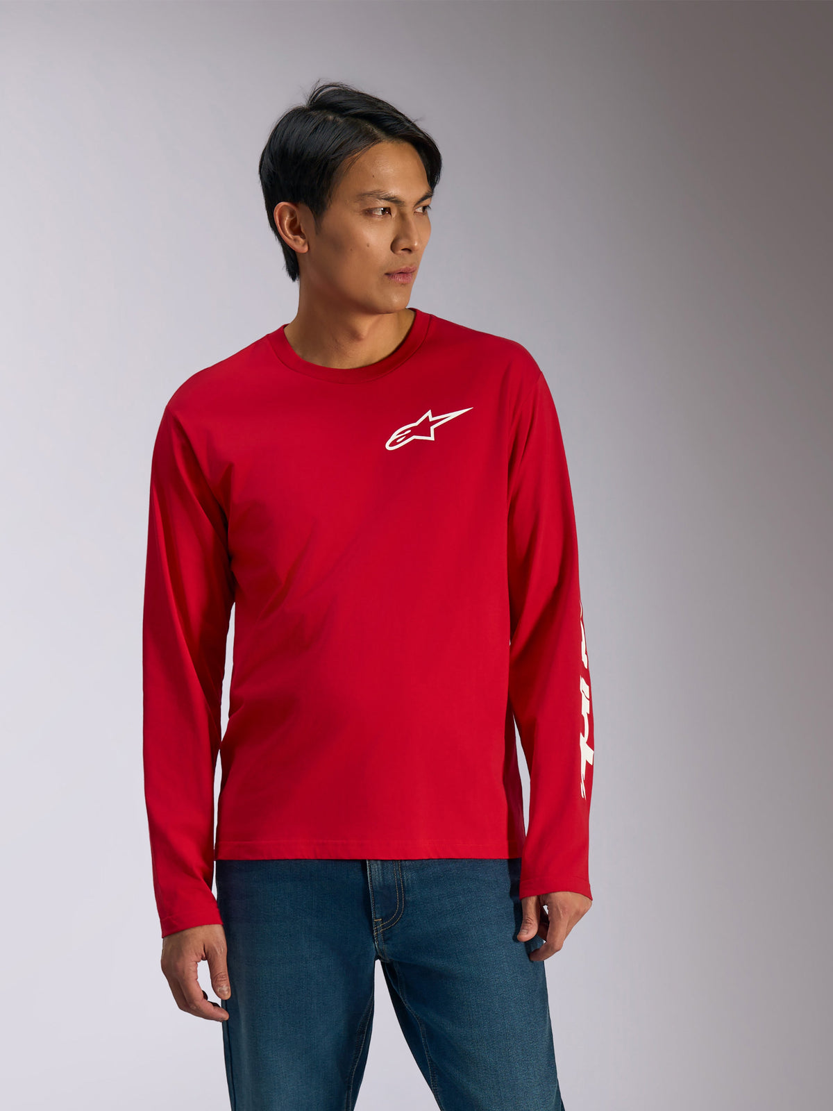 Alpinestars Trackside CSF Tee, koszulka z długim rękawem, czerwona z białymi grafikami, białe logo gwiazdy Alpinestars na piersi i biały napis na rękawie, noszona z niebieskimi dżinsami, codzienna odzież sportowa inspirowana motorsportem