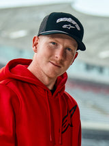 Man wearing a red Alpinestars hoodie and the Agree Trucker Hat in Black, featuring white embroidered Astars logos, a white braided rope detail on the visor, and a breathable mesh back, casual lifestyle setting