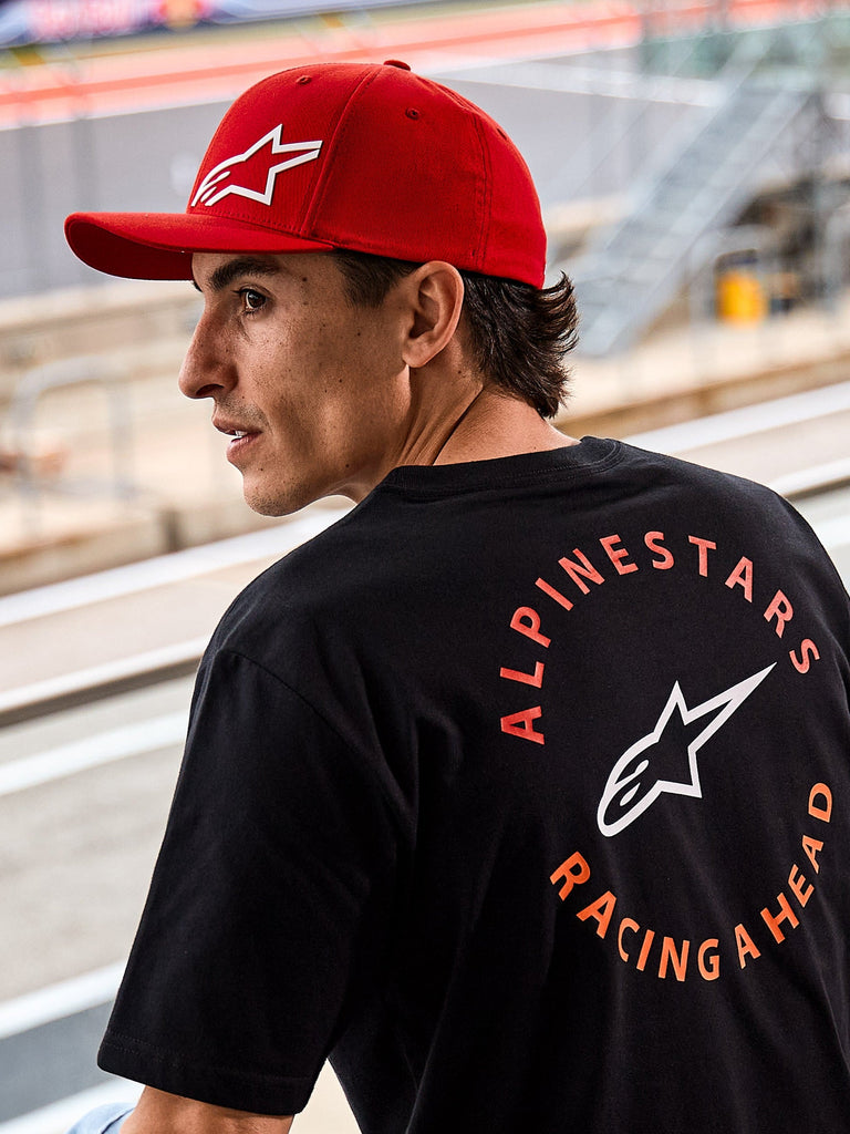 Alpinestars Corp Shift Multi Hat, czerwona czapka z zakrzywionym daszkiem i białym logo Astars 3D, noszona z czarnym t-shirtem Alpinestars z grafiką „Racing Ahead”, lifestyle'owa sceneria motorsportowa na torze wyścigowym