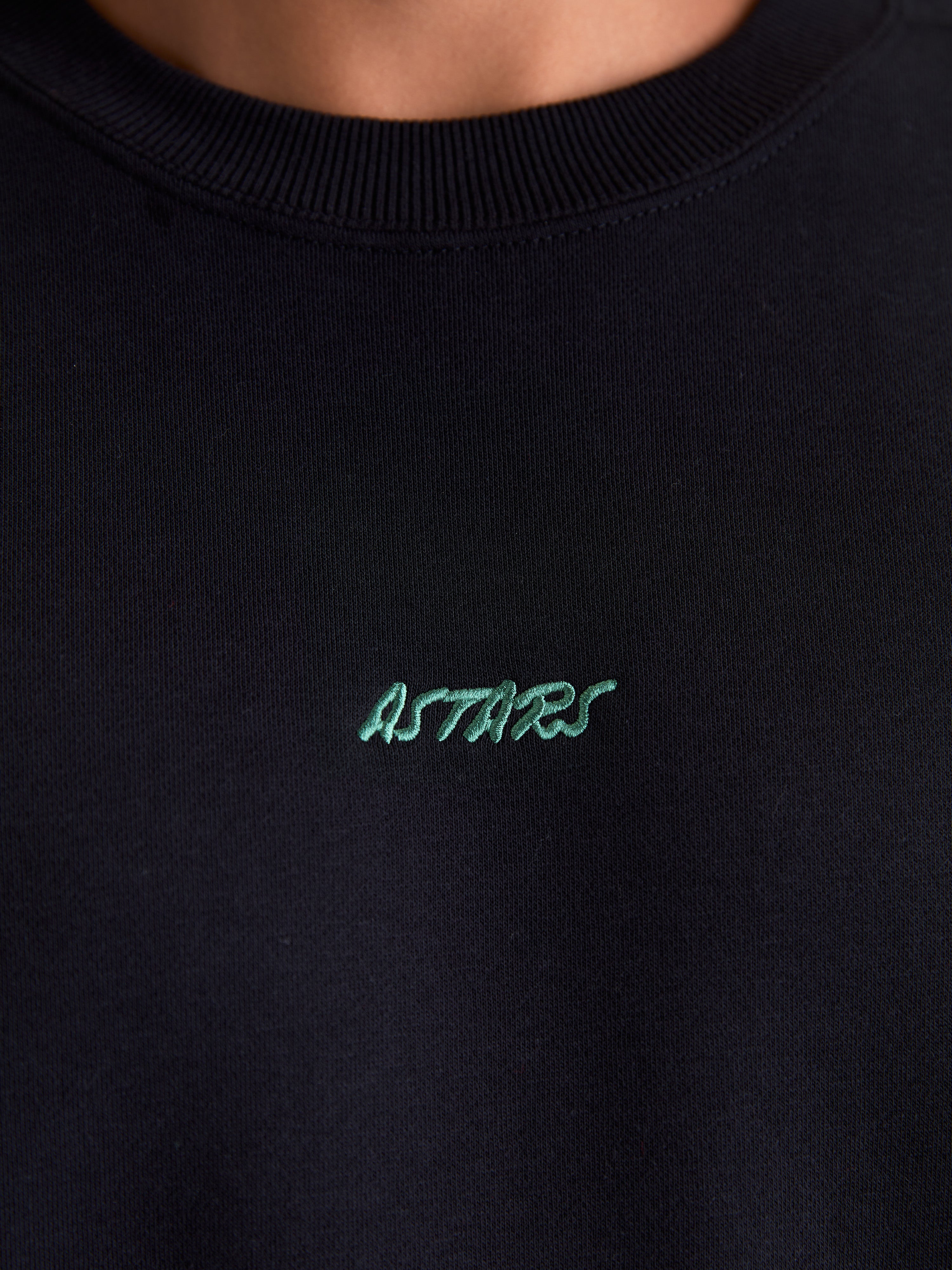 Alpinestars Guise Oversize, czarna sportowa bluza polarowa z okrągłym dekoltem, zbliżenie na prążkowany dekolt i zielone haftowane logo Astars na piersi, bawełna i poliester z recyklingu.