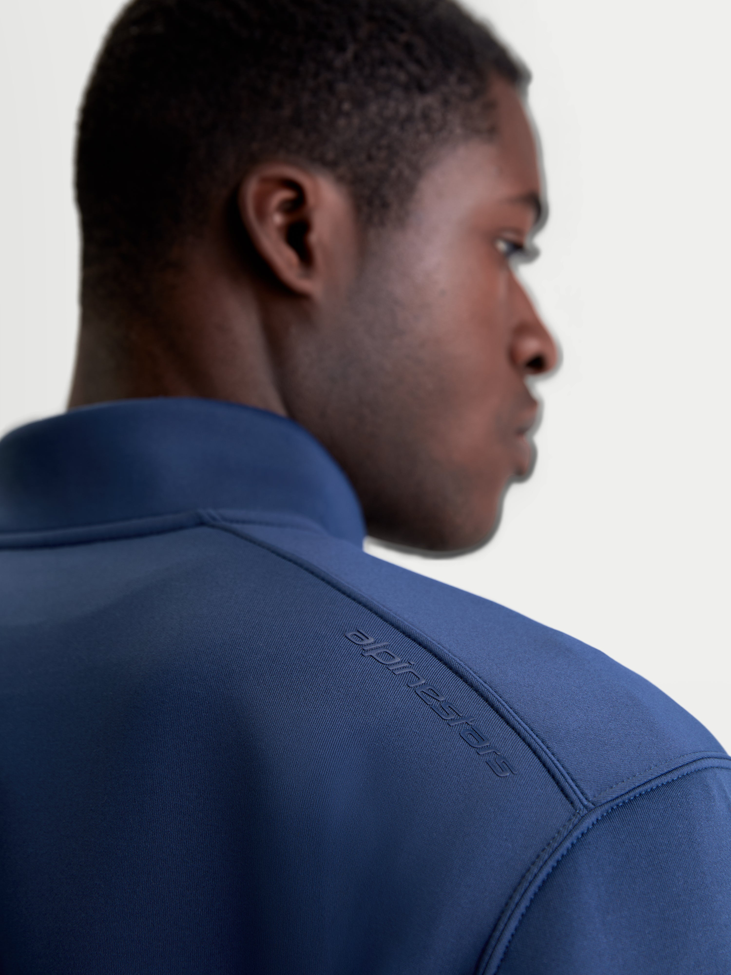 Alpinestars Bluza Apera Half-Zip, niebieska bluza sportowa typu pullover, zbliżenie na tył na modelu, tonalny nadruk logo HD na ramieniu, wykonana z materiału polarowego tri-blend premium, odzież sportowa w stylu motorsport.