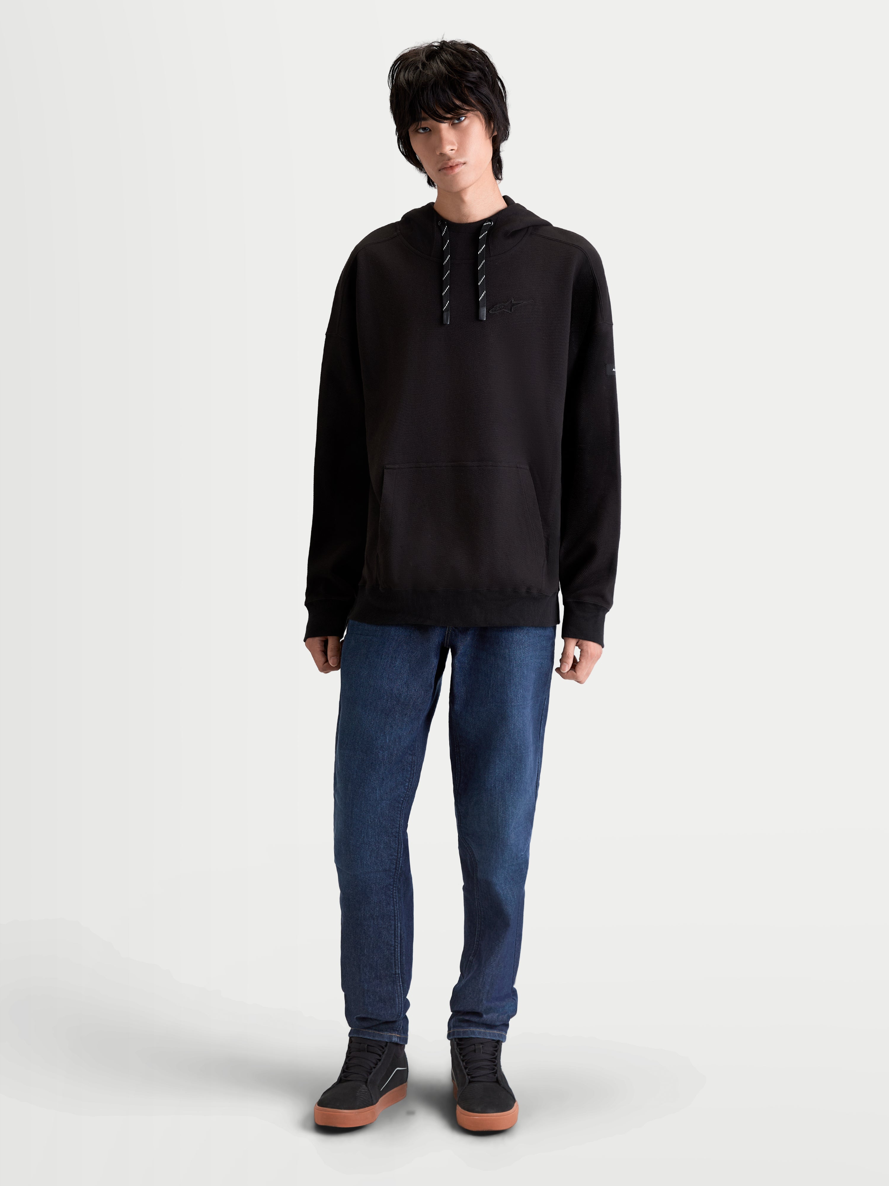 Alpinestars Bluza Z Kapturem Camino Textured Oversized, męska bluza lifestyle typu oversize, czarna, teksturowany polar o splocie twill, wysoki kołnierz i kieszeń typu kangurka, haftowane logo, model w niebieskich jeansach.