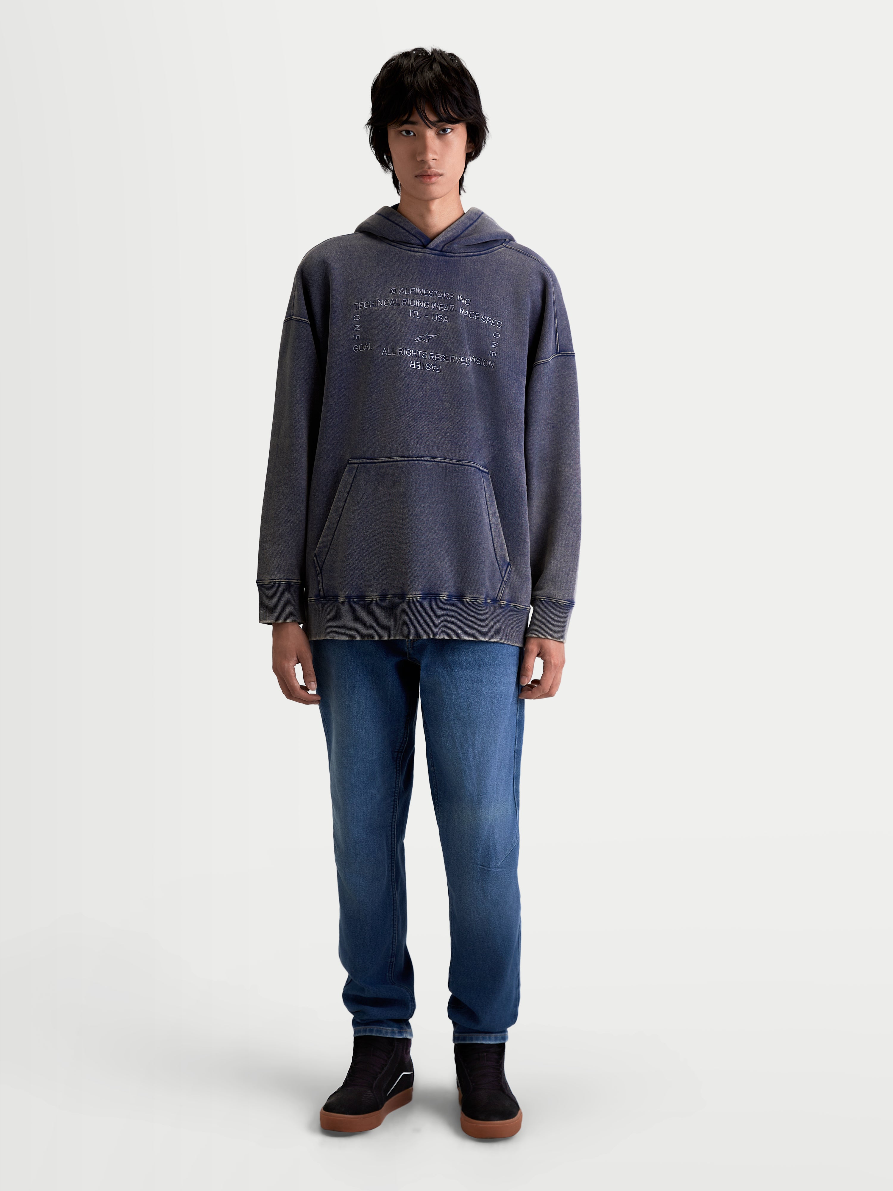 Alpinestars bluza z kapturem Infinite Washed Oversized, casualowa bluza sportowa typu pullover noszona przez modela, granatowa z wykończeniem snow-wash, kieszeń kangurka i haftowany napis Alpinestars na klatce piersiowej, bawełna premium i poliester z recyklingu, krój oversize zapewniający komfort.