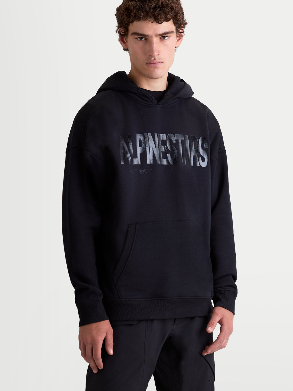 Alpinestars Bluza z kapturem Shaw Oversized, czarna męska bluza o luźnym kroju, teksturowane logo na klatce piersiowej, kieszeń typu kangurka, kaptur z podszewką, polar z bawełny i poliestru z recyklingu, widok z przodu na modelu