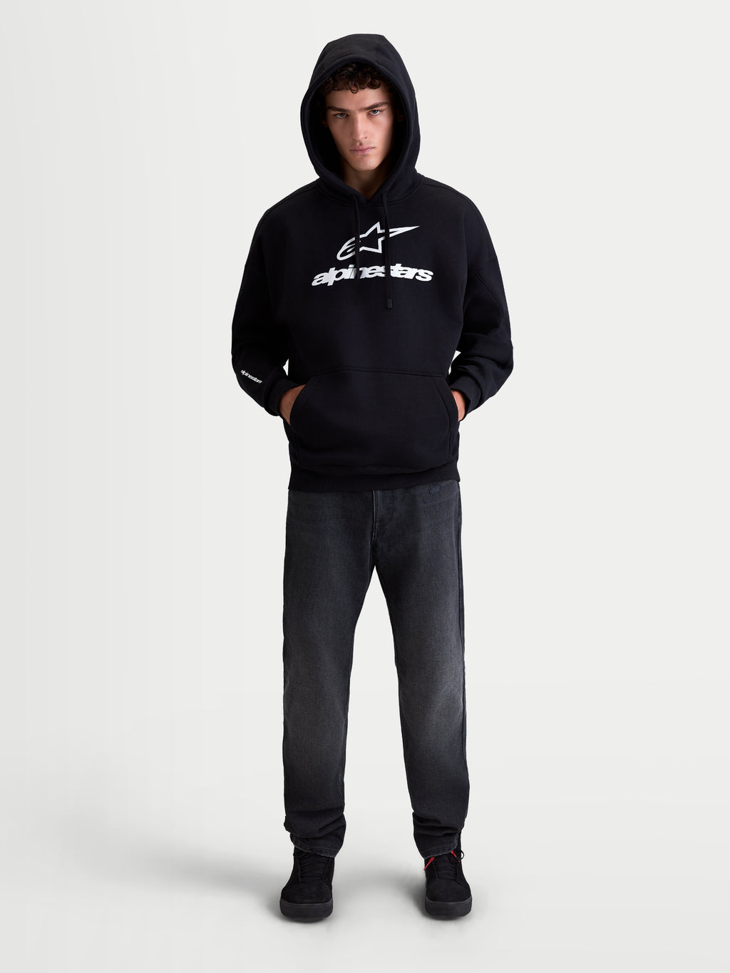 Alpinestars Bluza Z Kapturem Prelude Oversized, sportowa bluza polarowa typu pullover, unisex, czarna i biała, bawełna i poliester z recyklingu, luźny fason oversized z kieszenią kangurka i wyrazistym, wypukłym logo na klatce piersiowej i rękawie, pokazana na modelu.