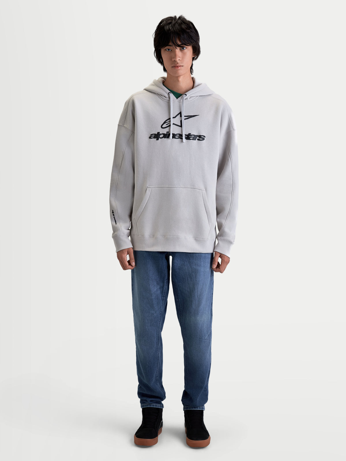 Alpinestars bluza z kapturem Prelude Oversize, sportowa bluza polarowa noszona przez modela, szary kolor z czarnym wypukłym logo, luźny fason oversize, kieszeń kangurka, kaptur z regulacją, mankiety ze ściągaczami, wykonana z bawełny i poliestru z recyklingu.