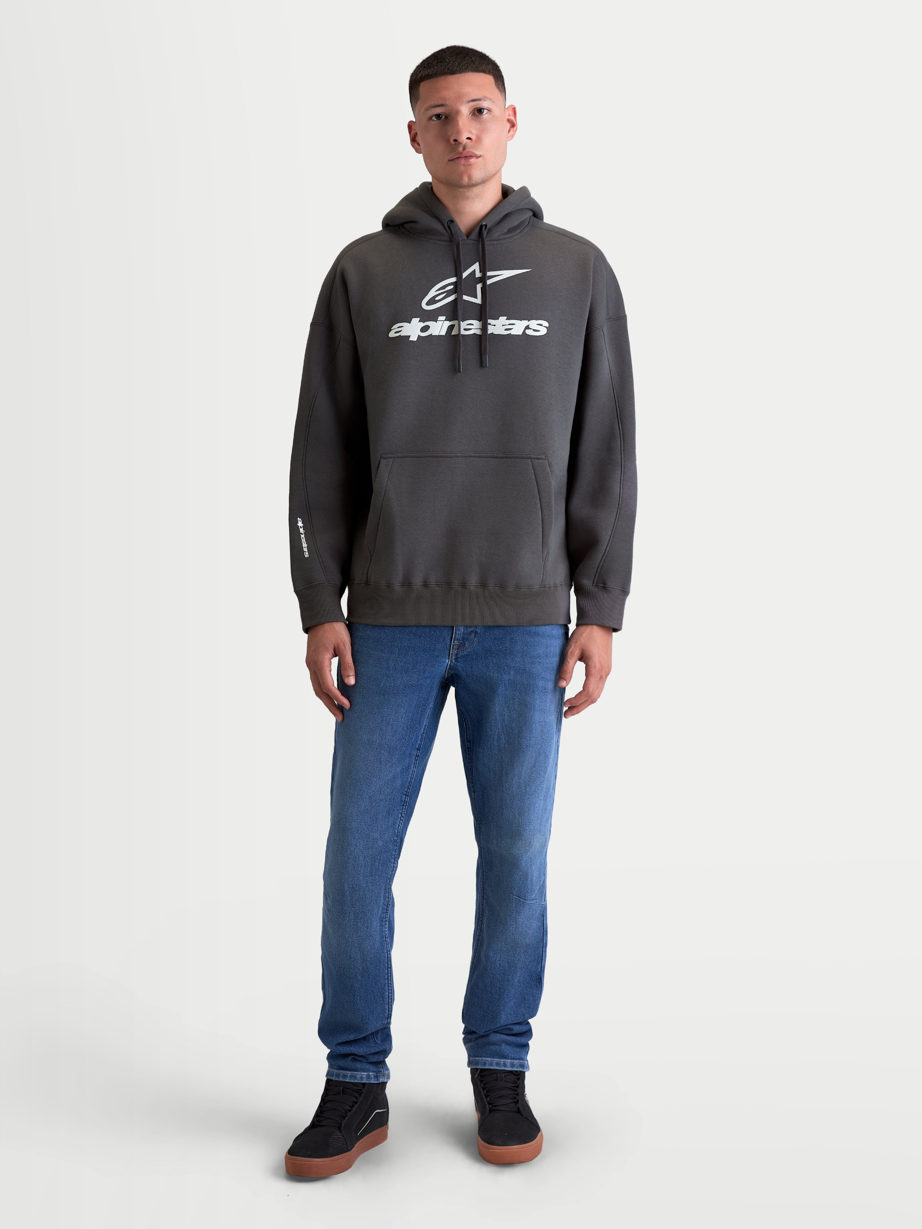 Alpinestars Prelude Oversize Hoodie, szara sportowa bluza z kapturem, luźny fason oversize, kieszeń kangurka, wypukłe nadruki logo na piersi i rękawie, polarowa odzież sportowa dla mężczyzn i kobiet, prezentowana na modelu.