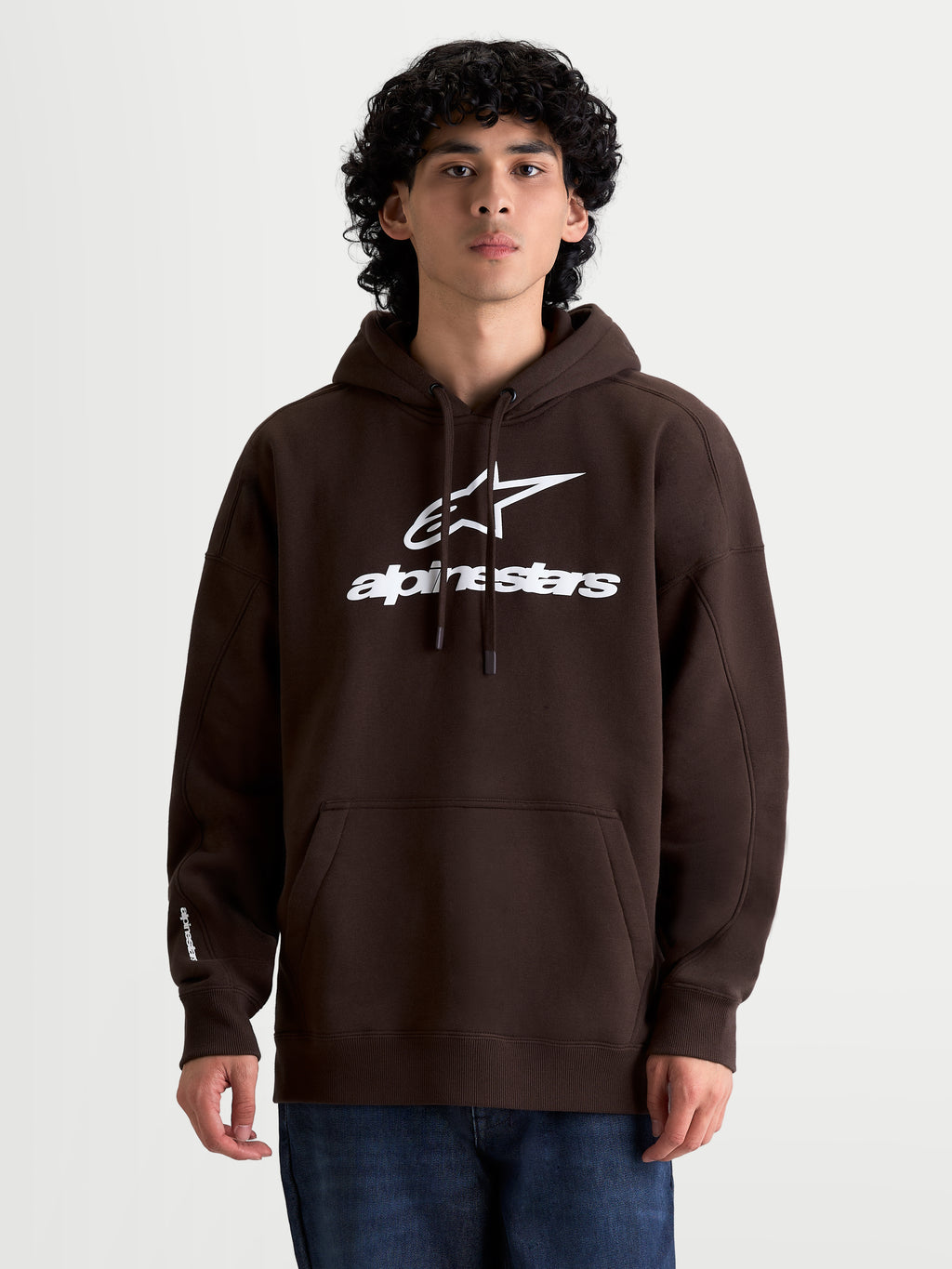 Alpinestars bluza z kapturem Prelude Oversized, brązowa męska bluza z kapturem na modelu, białe wypukłe logo Alpinestars, luźny krój oversized, kieszeń kangurka, kaptur ze sznurkiem, sportowa odzież codzienna.