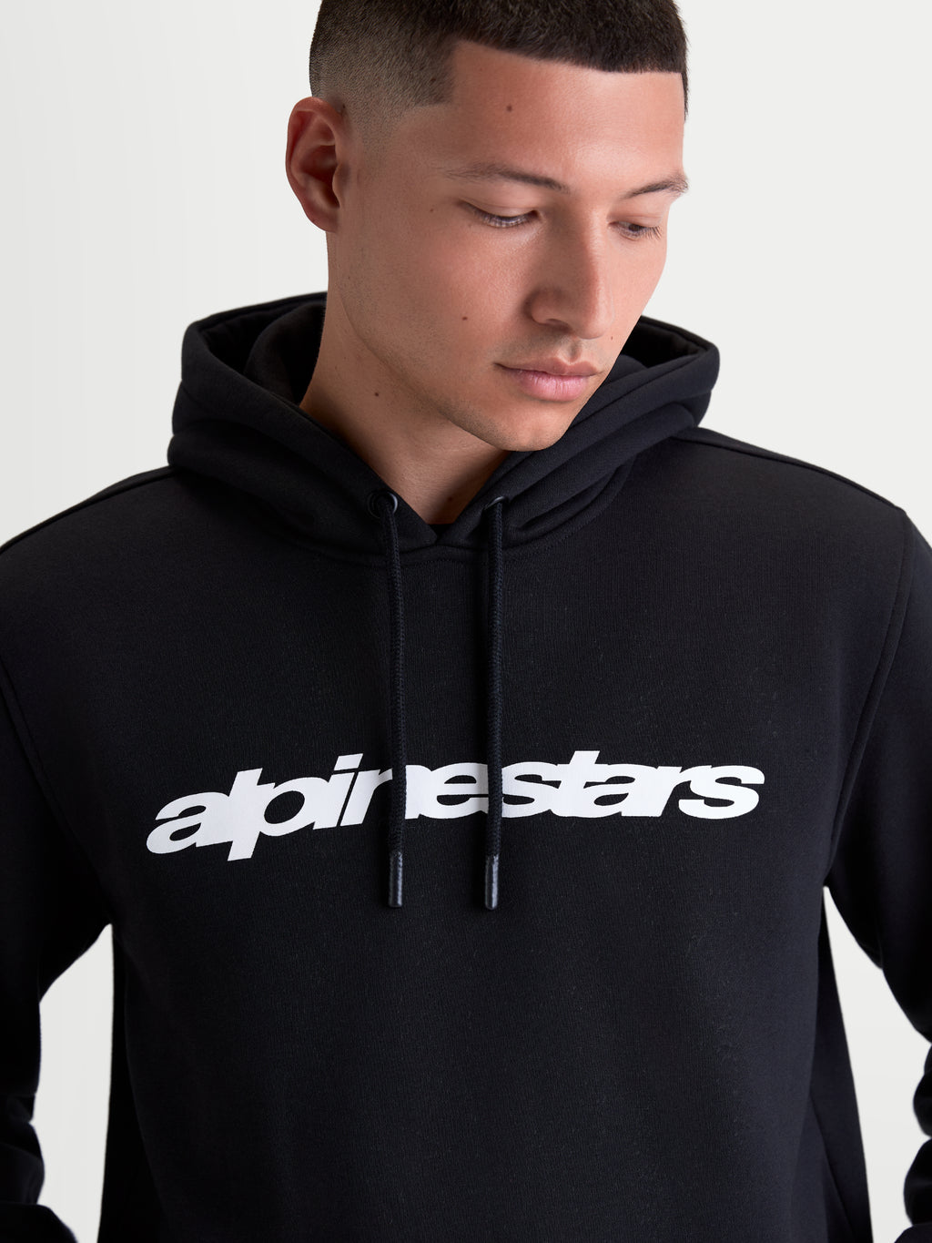 Alpinestars bluza z kapturem Race Wordmark, męska bluza z kapturem typu fleece, czarna i biała, wyrazisty biały logotyp wordmark na piersi, regulowany kaptur z podszewką i troczkami, odzież typu sportswear inspirowana motorsportem, wykonana z bawełny i poliestru z recyklingu, model prezentujący produkt.