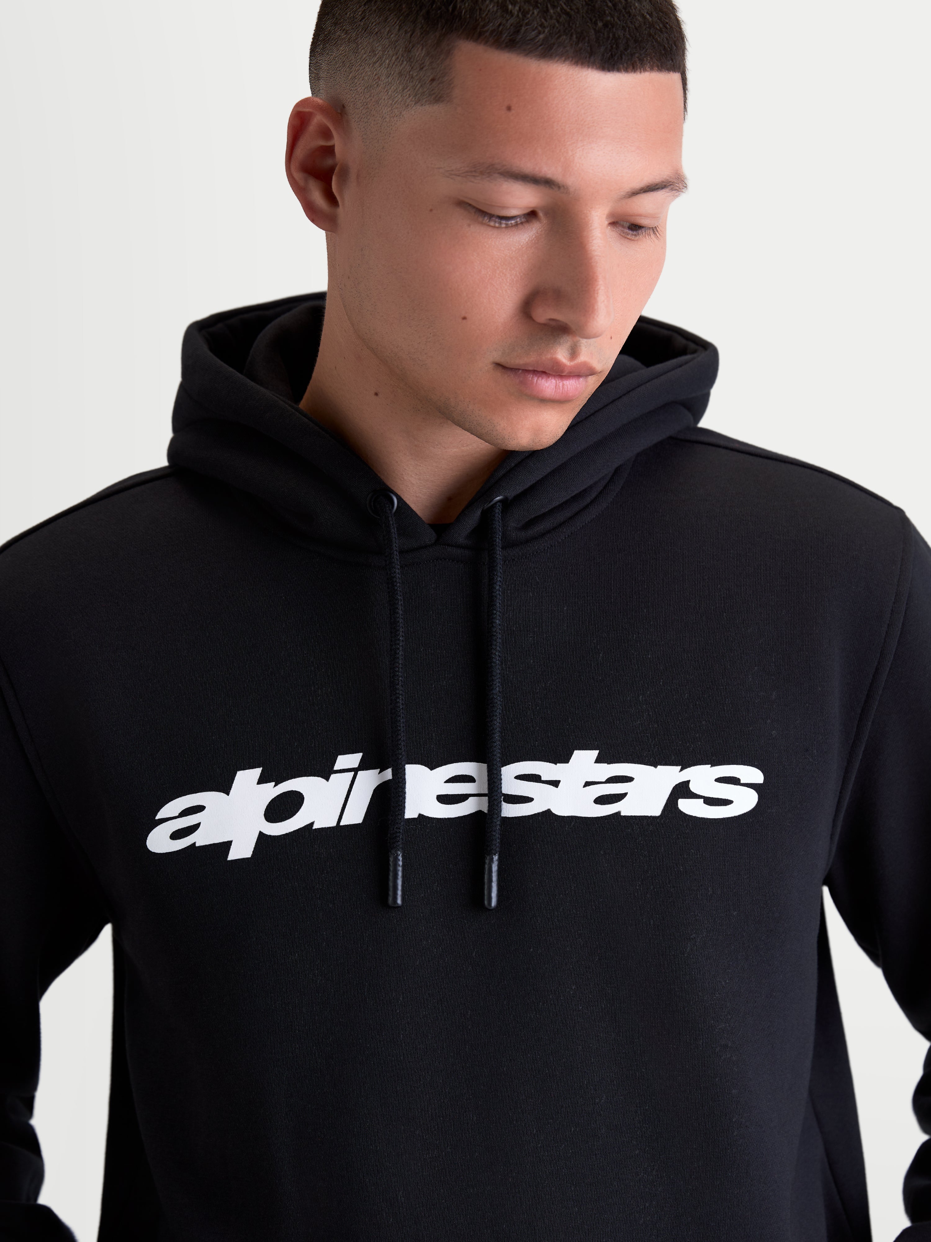 Alpinestars bluza z kapturem Race Wordmark, męska bluza z kapturem typu fleece, czarna i biała, wyrazisty biały logotyp wordmark na piersi, regulowany kaptur z podszewką i troczkami, odzież typu sportswear inspirowana motorsportem, wykonana z bawełny i poliestru z recyklingu, model prezentujący produkt.