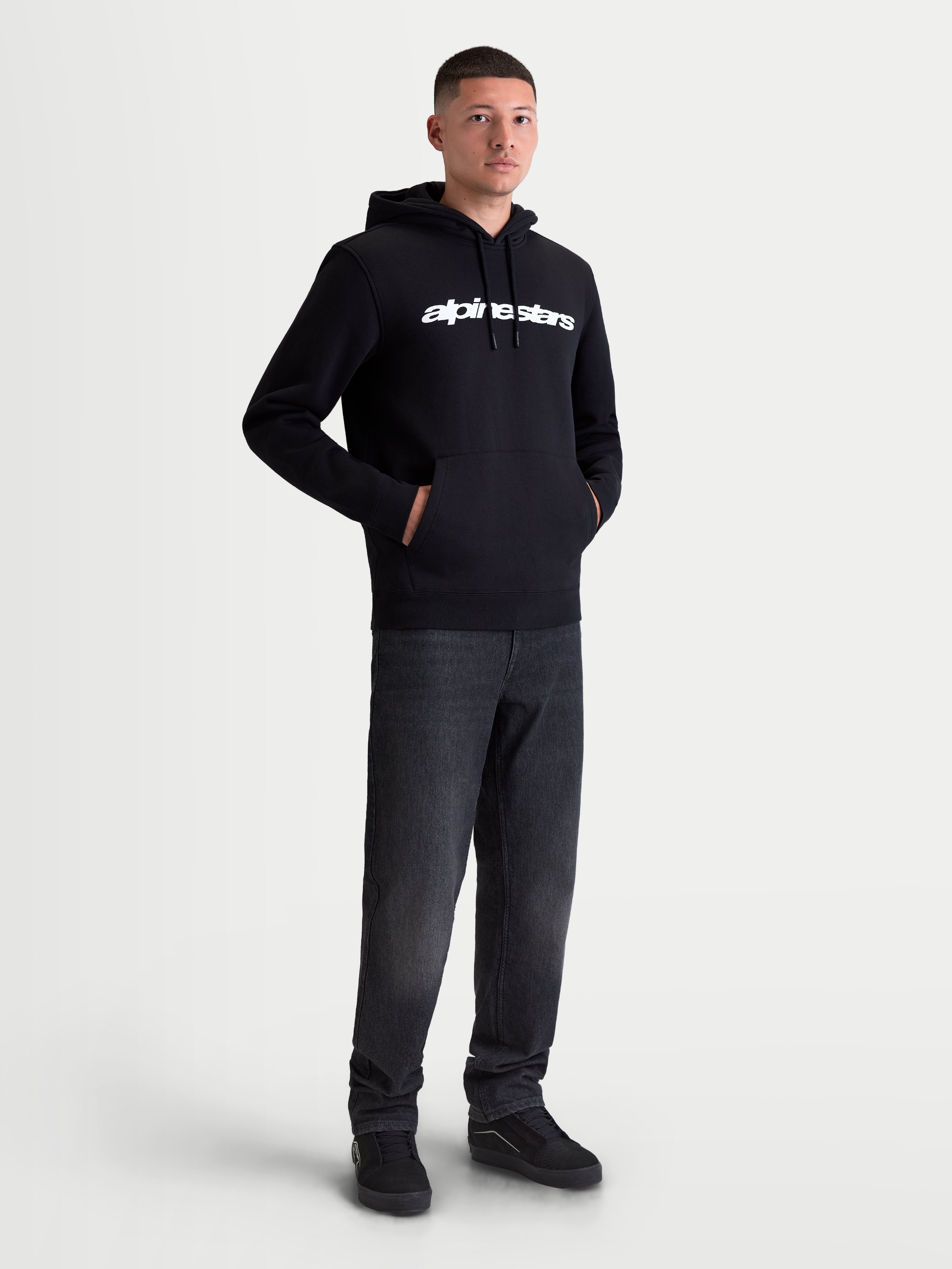 Alpinestars bluza z kapturem Race Wordmark, męska bluza dresowa, czarna, biały linearny logotyp na klatce piersiowej, kaptur z podszewką i sznurkami, przednia kieszeń typu kangurka, casualowa odzież sportowa w stylu motorsport, prezentowana na modelu.