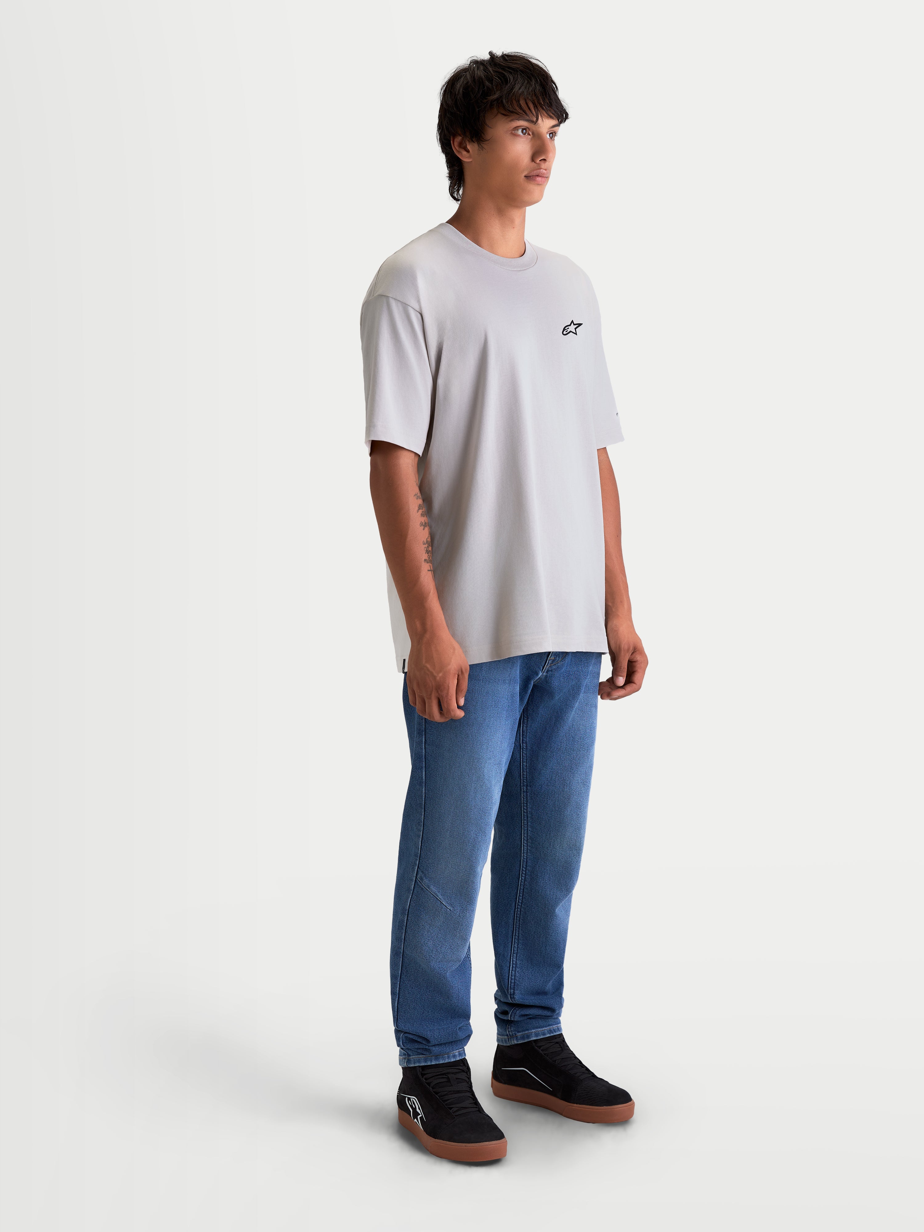 Alpinestars Mandate Oversized CSF Tee, szara koszulka z krótkim rękawem o kroju oversize, małe czarne logo Astars na piersi, casualowa odzież sportowa, model męski w niebieskich jeansach i czarnych butach.