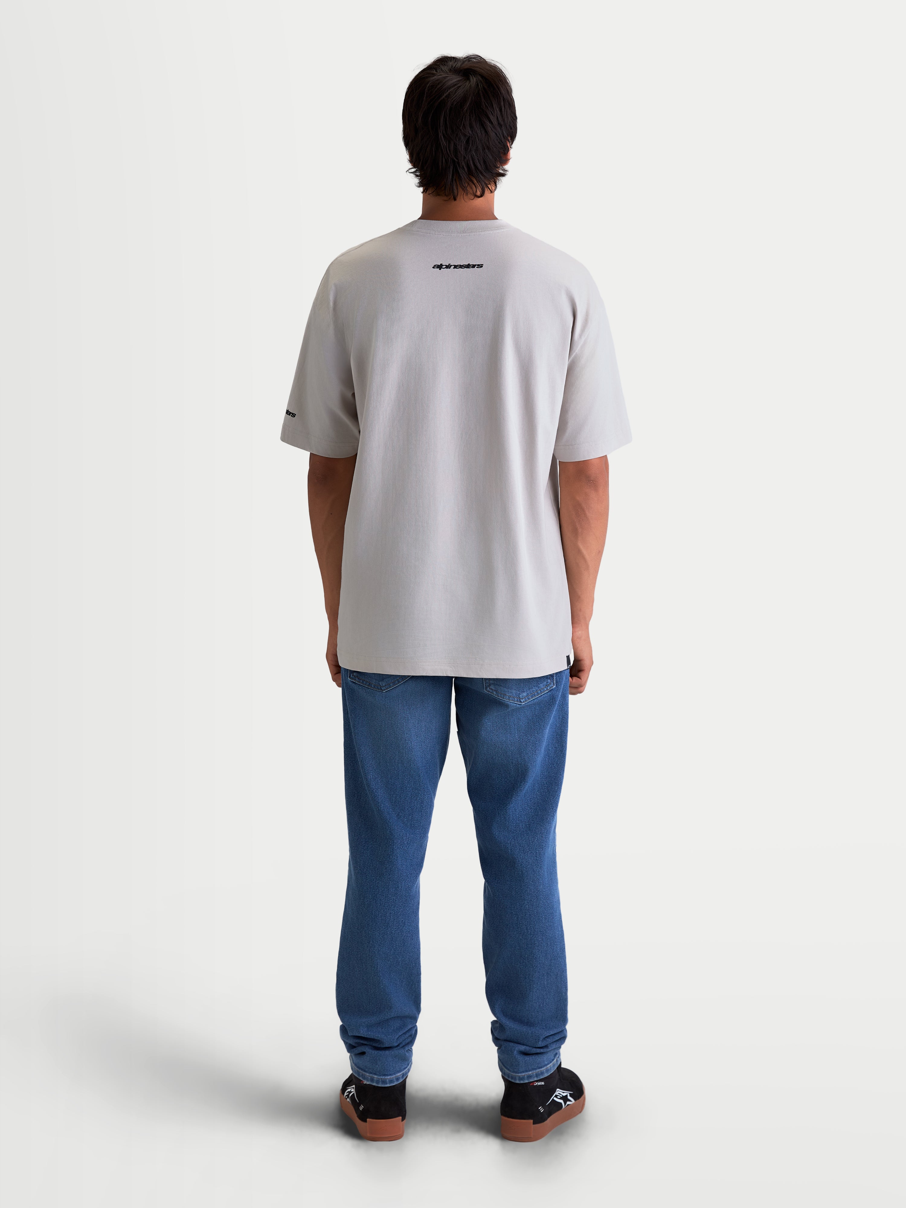 Alpinestars Mandate Oversized CSF, koszulka z krótkim rękawem, sportswear, szary cementowy, widok z tyłu na modelu. Koszulka o kroju oversize z czarnym napisem Alpinestars na plecach, wykonana z miękkiej mieszanki bawełny i poliestru.