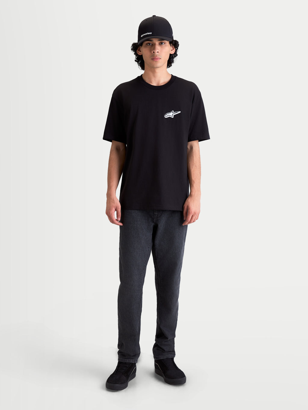 Alpinestars Koszulka Meteor CSF, męski t-shirt z krótkim rękawem, czarny z białym logo Astars na piersi, casualowa odzież sportowa inspirowana motorsportem, model ma na sobie ciemne spodnie i czapkę z daszkiem.