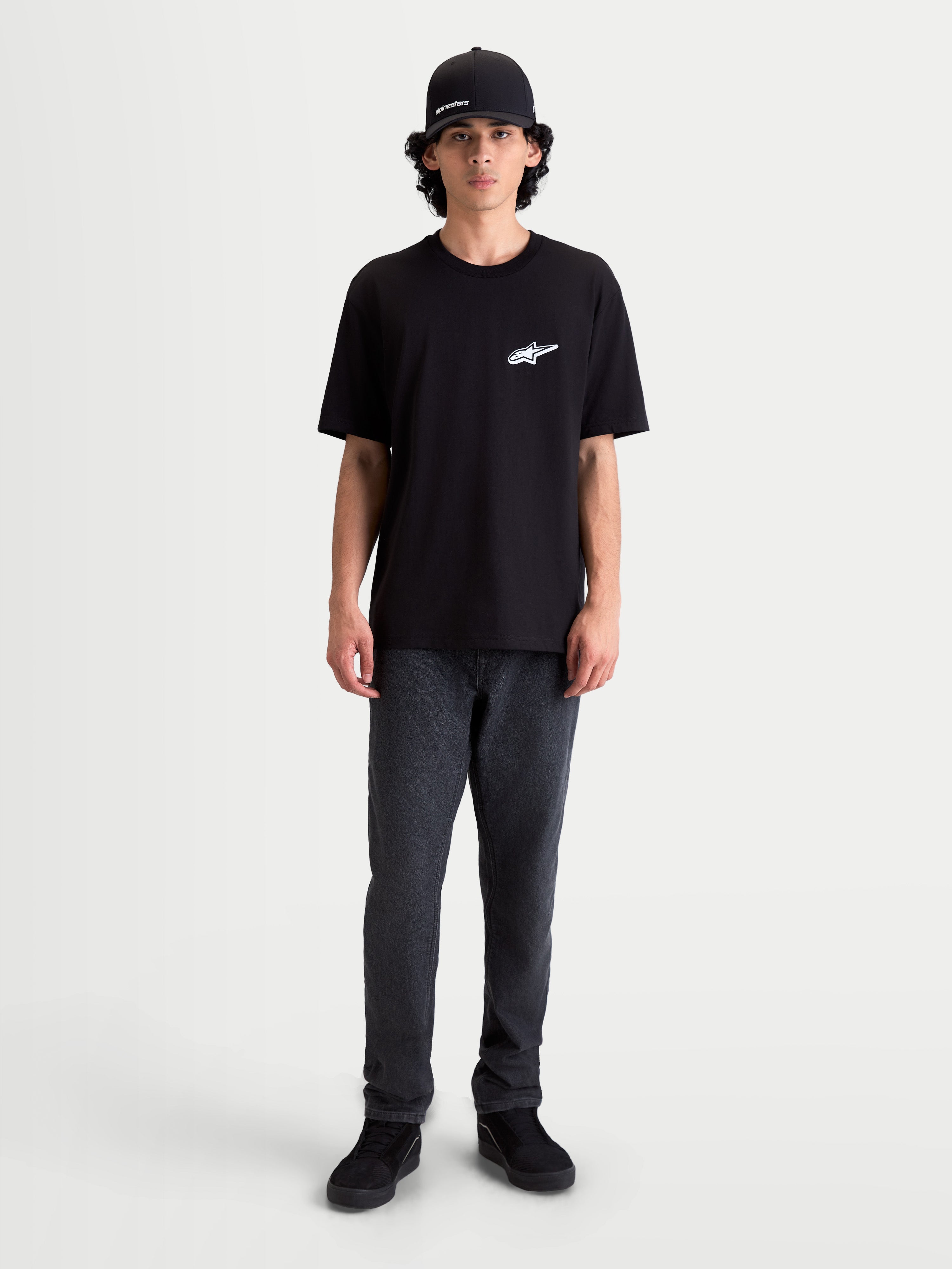 Alpinestars Koszulka Meteor CSF, męski t-shirt z krótkim rękawem, czarny z białym logo Astars na piersi, casualowa odzież sportowa inspirowana motorsportem, model ma na sobie ciemne spodnie i czapkę z daszkiem.