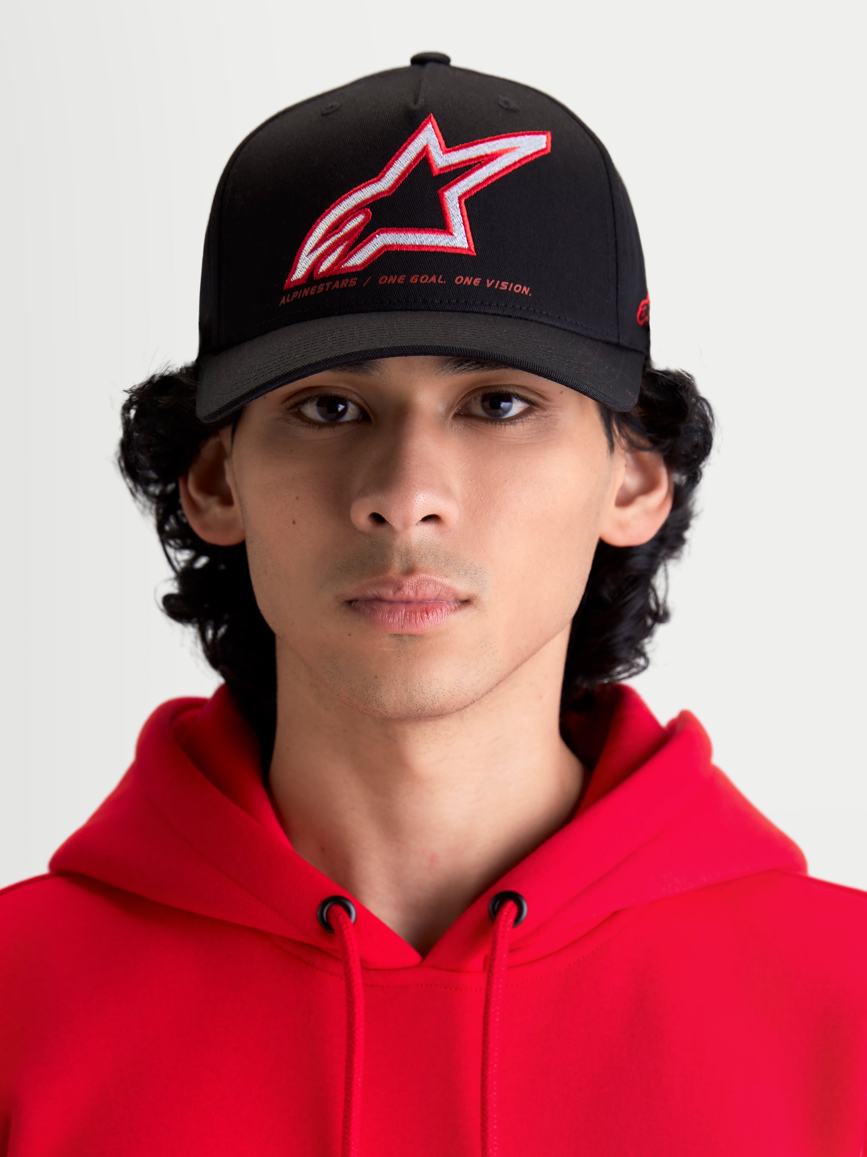 Alpinestars czapka Lineout, czarna czapka z daszkiem typu baseball, zakrzywiony daszek, czerwone i białe haftowane logo Astars oraz branding One Goal One Vision, widok z przodu na modelu w czerwonej bluzie z kapturem, usztywniona konstrukcja z twillu, odzież sportowa.